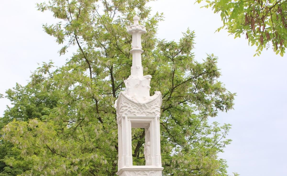 White stone column with cross in front of a green tree.