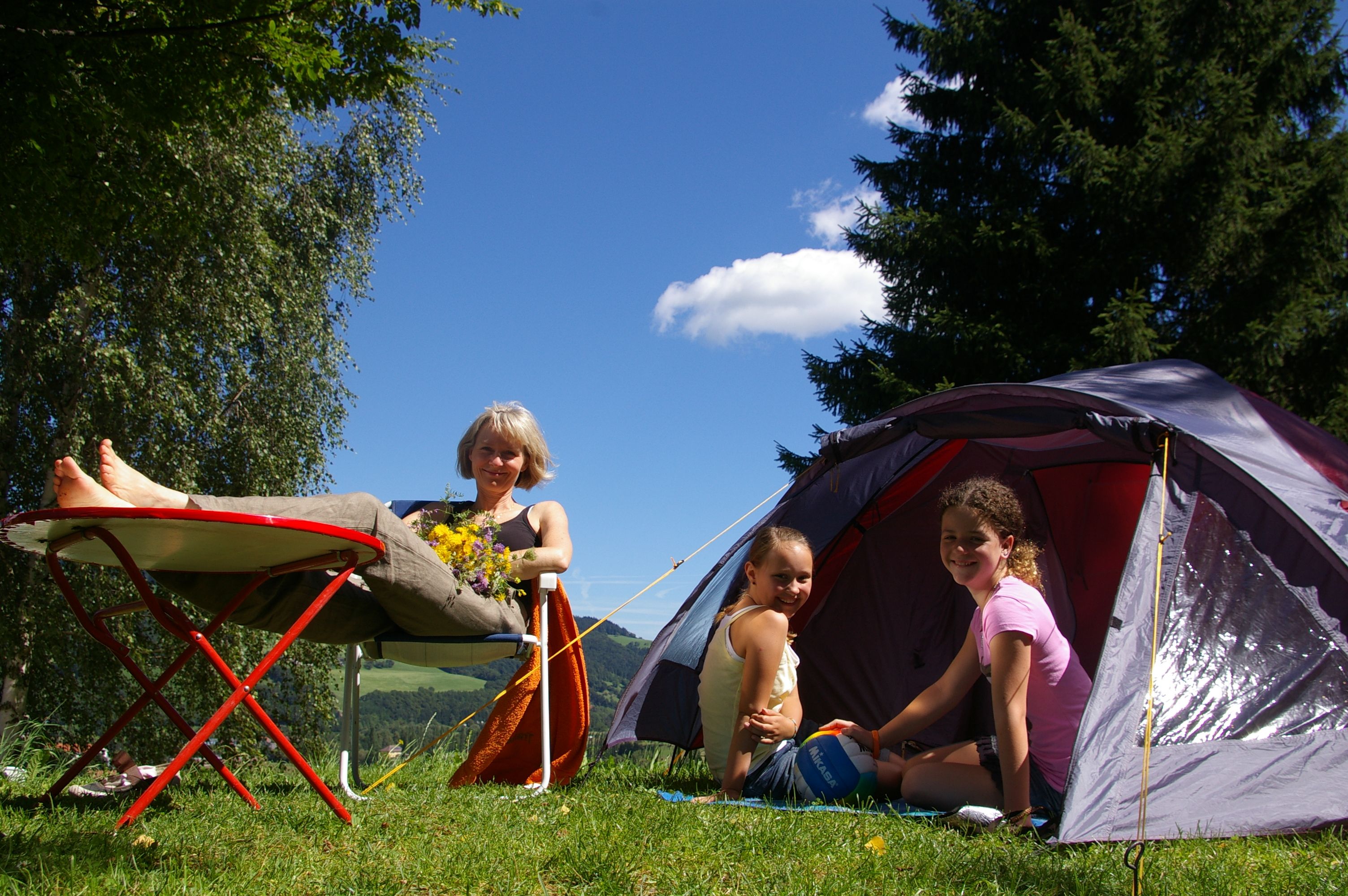 A woman relaxes on a camping chair next to a tent, while two children sit in the tent. Trees and blue sky can be seen in the background.