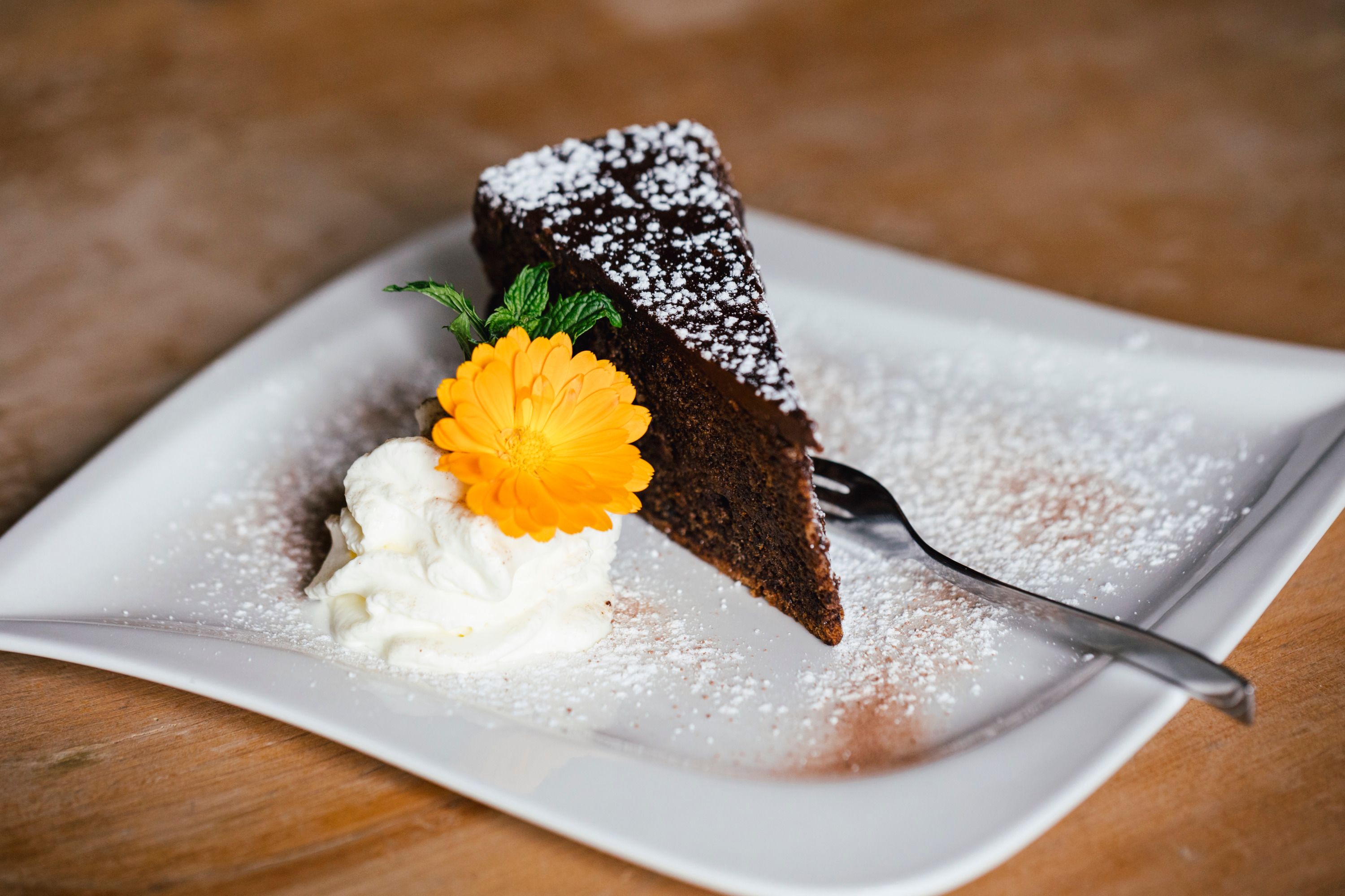 A piece of chocolate cake with powdered sugar, cream and a yellow flower on a plate.
