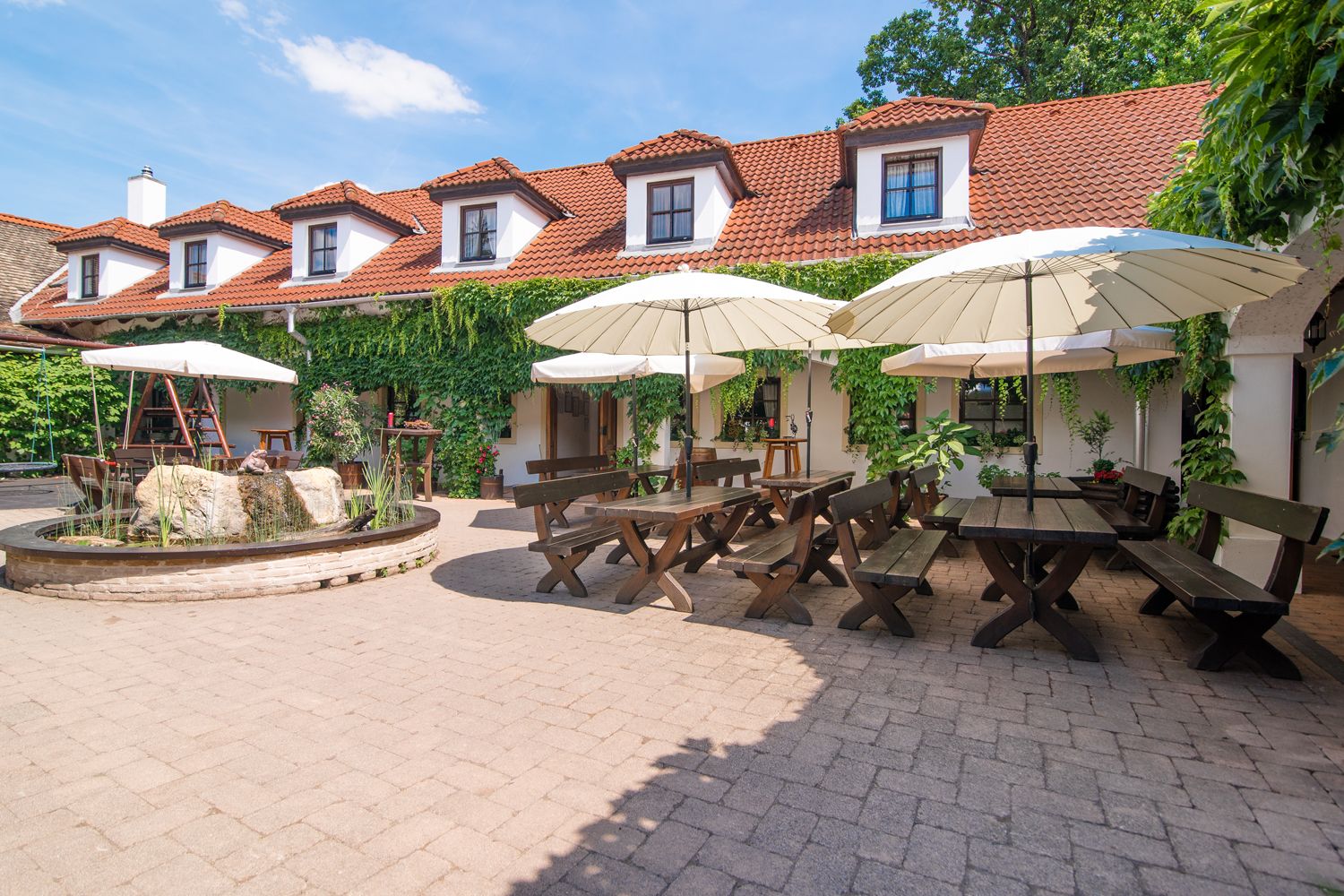 A cozy courtyard of a winery with wooden tables, benches and parasols, surrounded by a building with red tiled roofs and green vegetation.