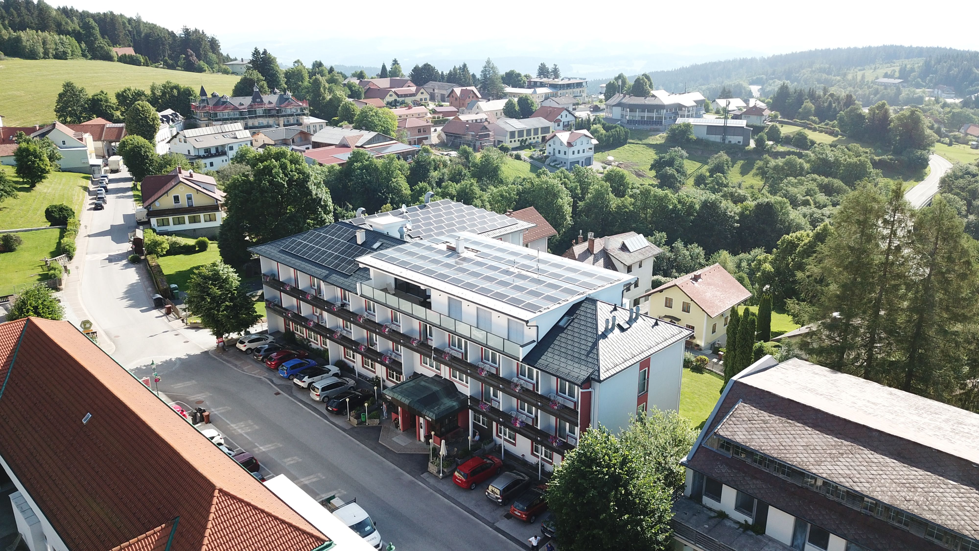 Aerial view of a hotel with solar panels on the roof, surrounded by trees and other buildings in a rural area.