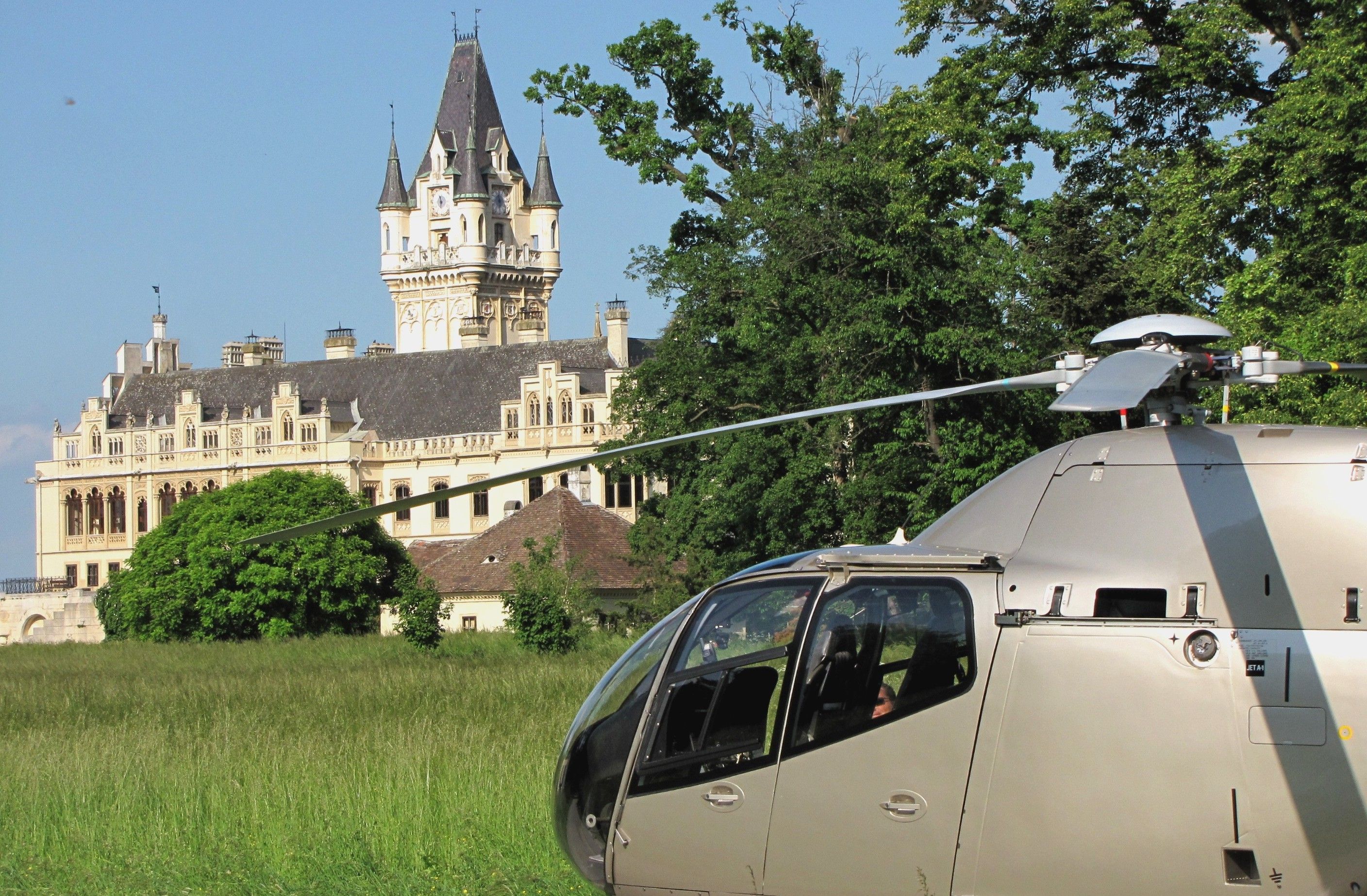 A helicopter stands on a meadow in front of a large, historic castle with towers and a clock tower.