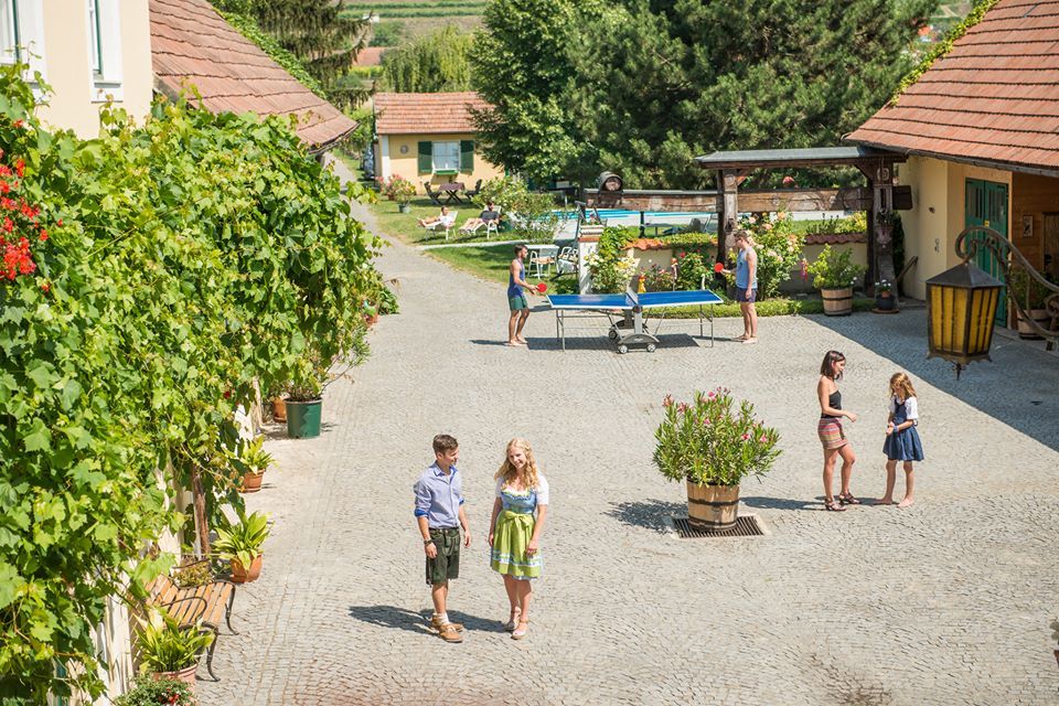 A sunny courtyard with people walking, playing table tennis and chatting. Surrounded by plants and buildings with red roofs.