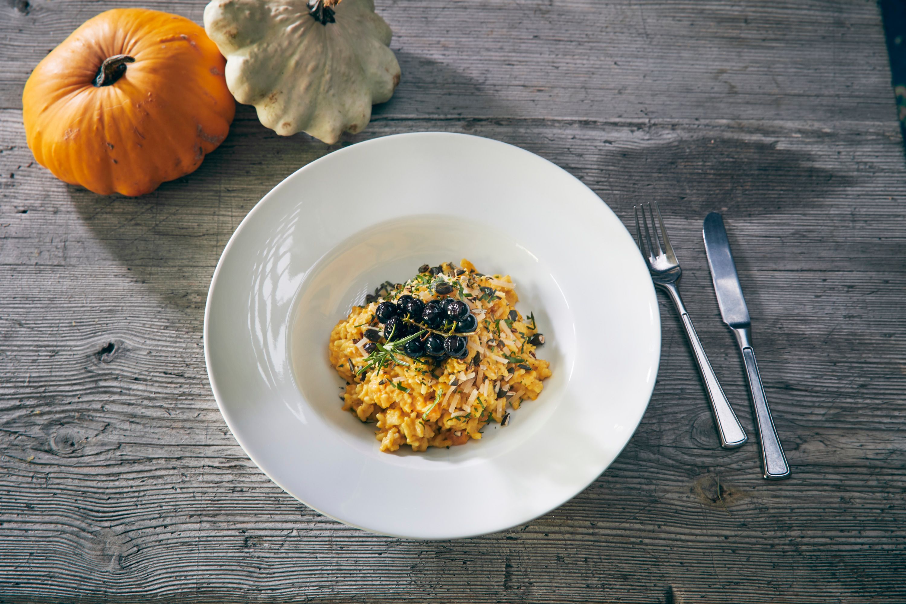 A plate of pumpkin risotto with herbs and berries, next to two pumpkins on a wooden table.