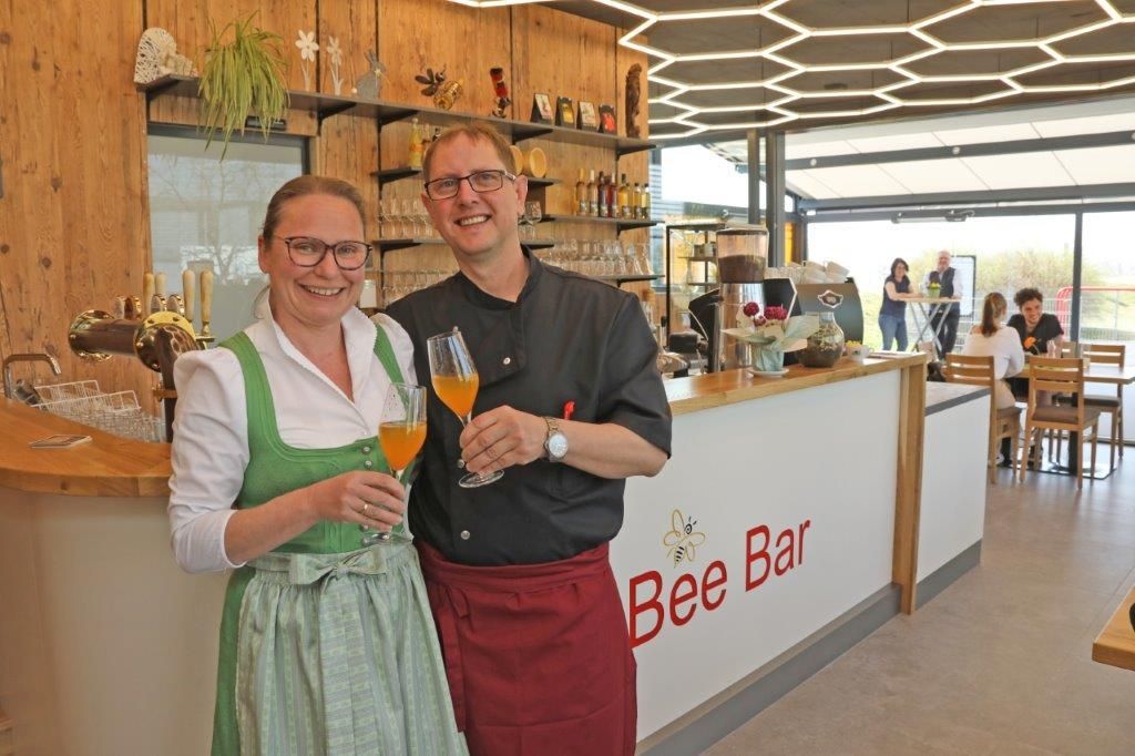 Two people in traditional dress stand smiling in front of the Bee Bar with drinks in their hands.