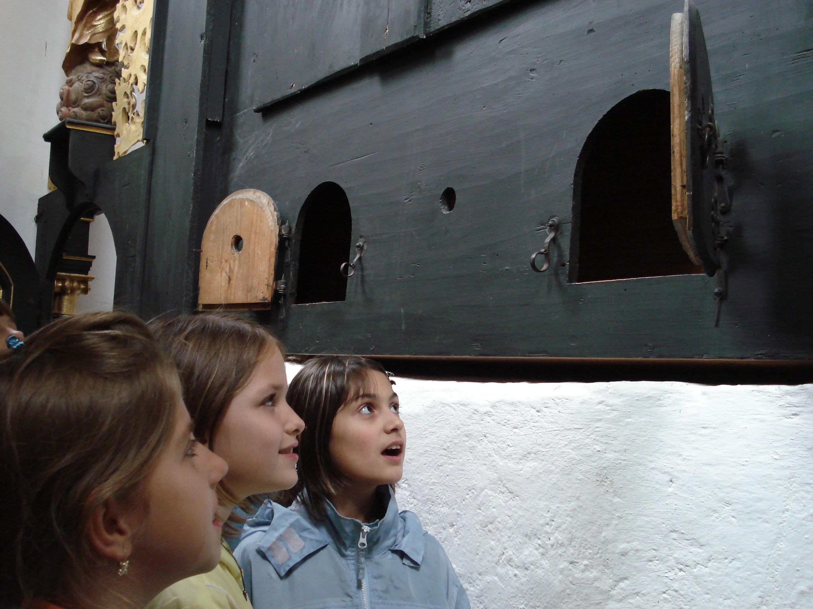 Three children look curiously at a wall with small doors.