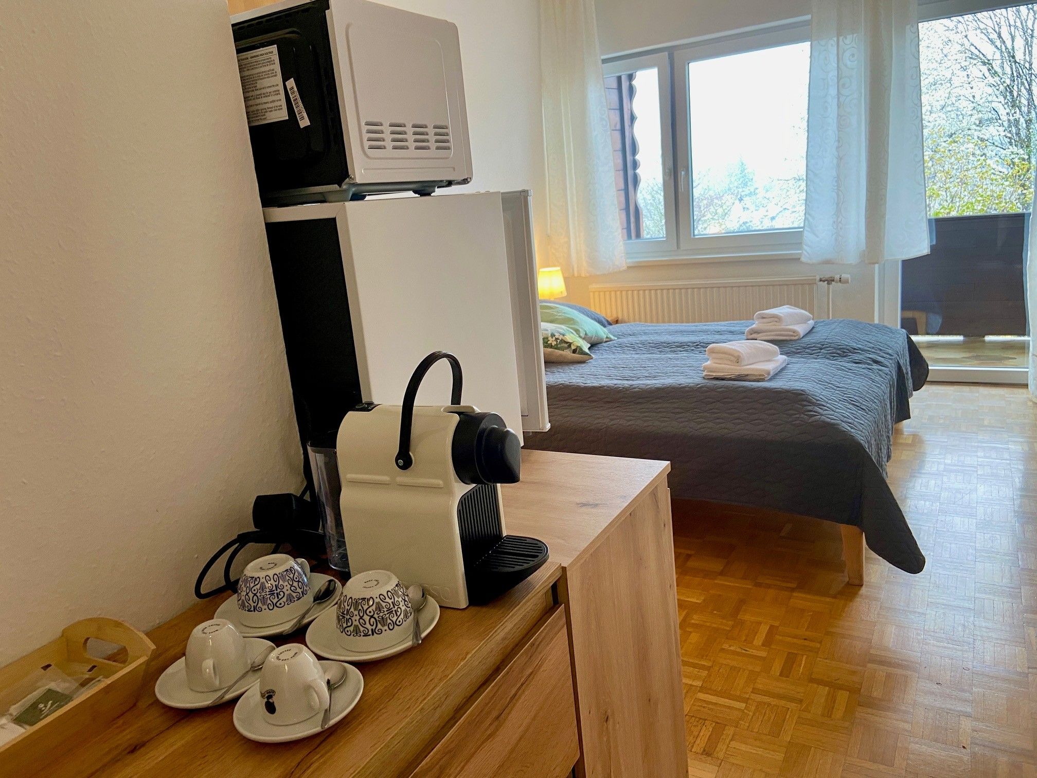 A hotel room with a coffee machine on a table and cups next to it. In the background a bed with towels and a window with a view outside.