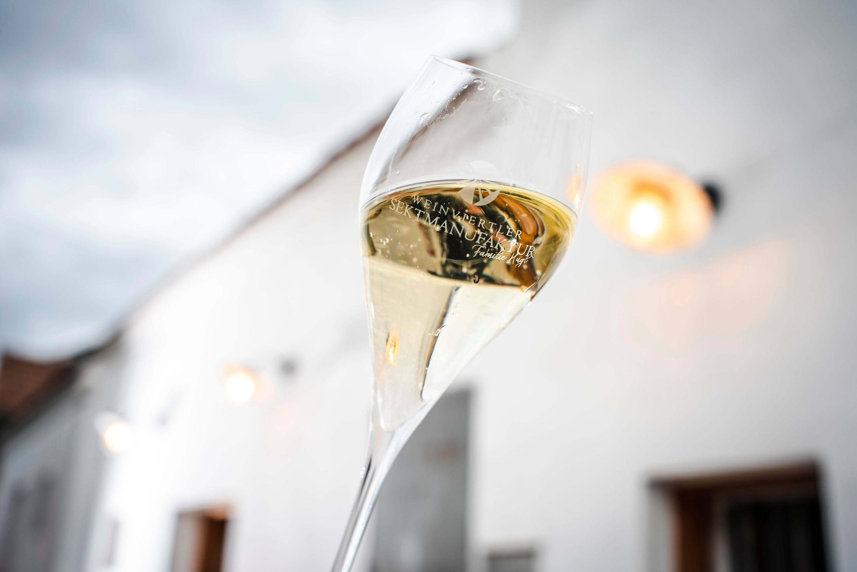 A glass of sparkling wine labeled 'Weinviertler Sektmanufaktur' is held against a cloudy sky.