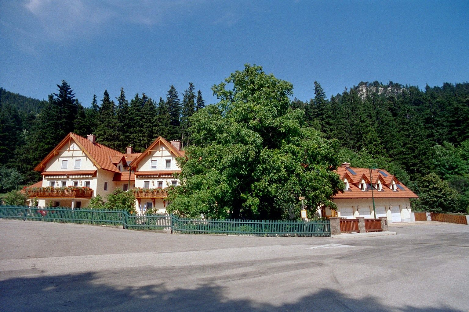 A large house with red roofs and balconies, surrounded by trees and a forest in the background.