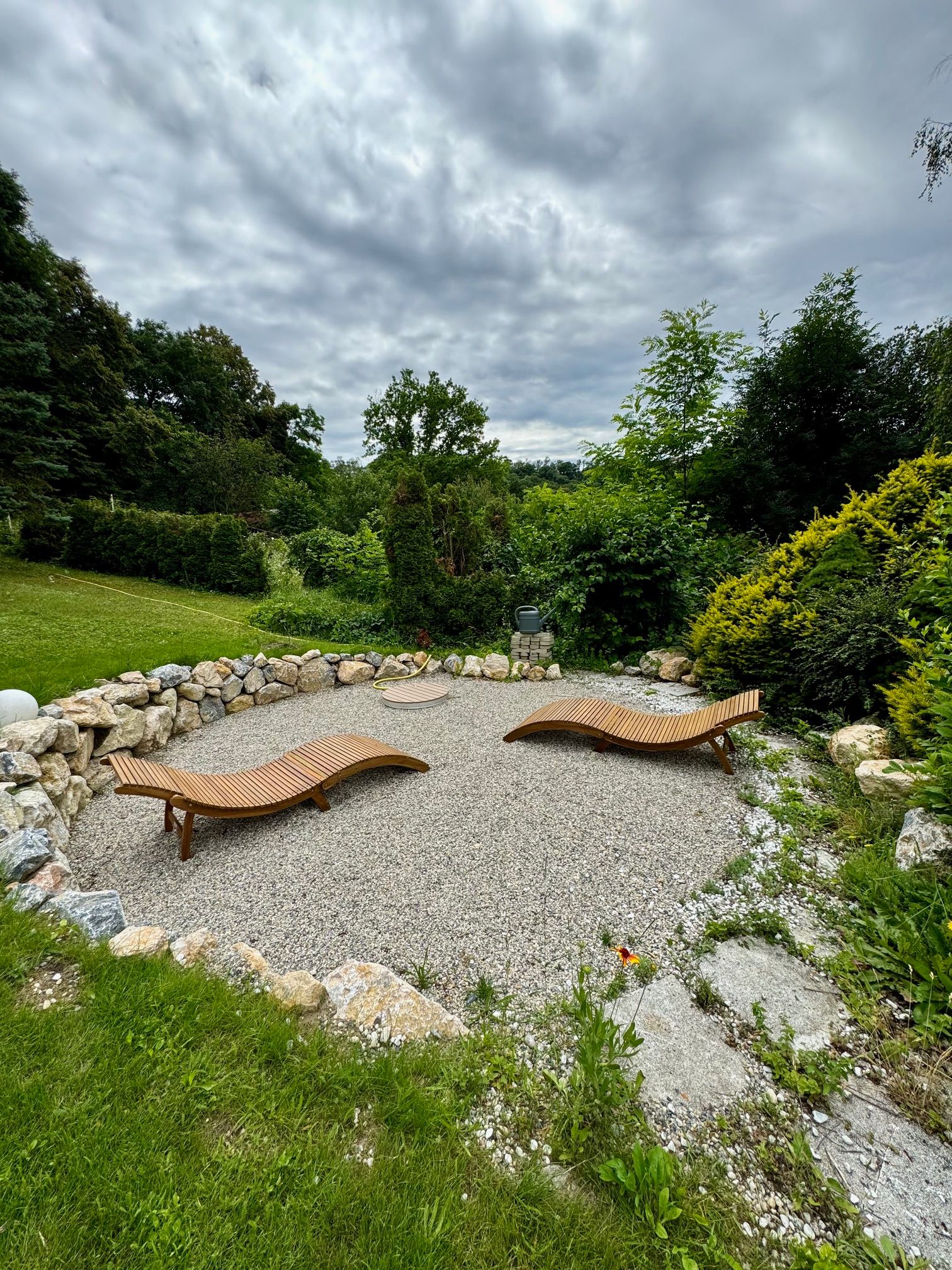 Two wooden loungers on a gravelled area, surrounded by stones and bushes, under a cloudy sky.