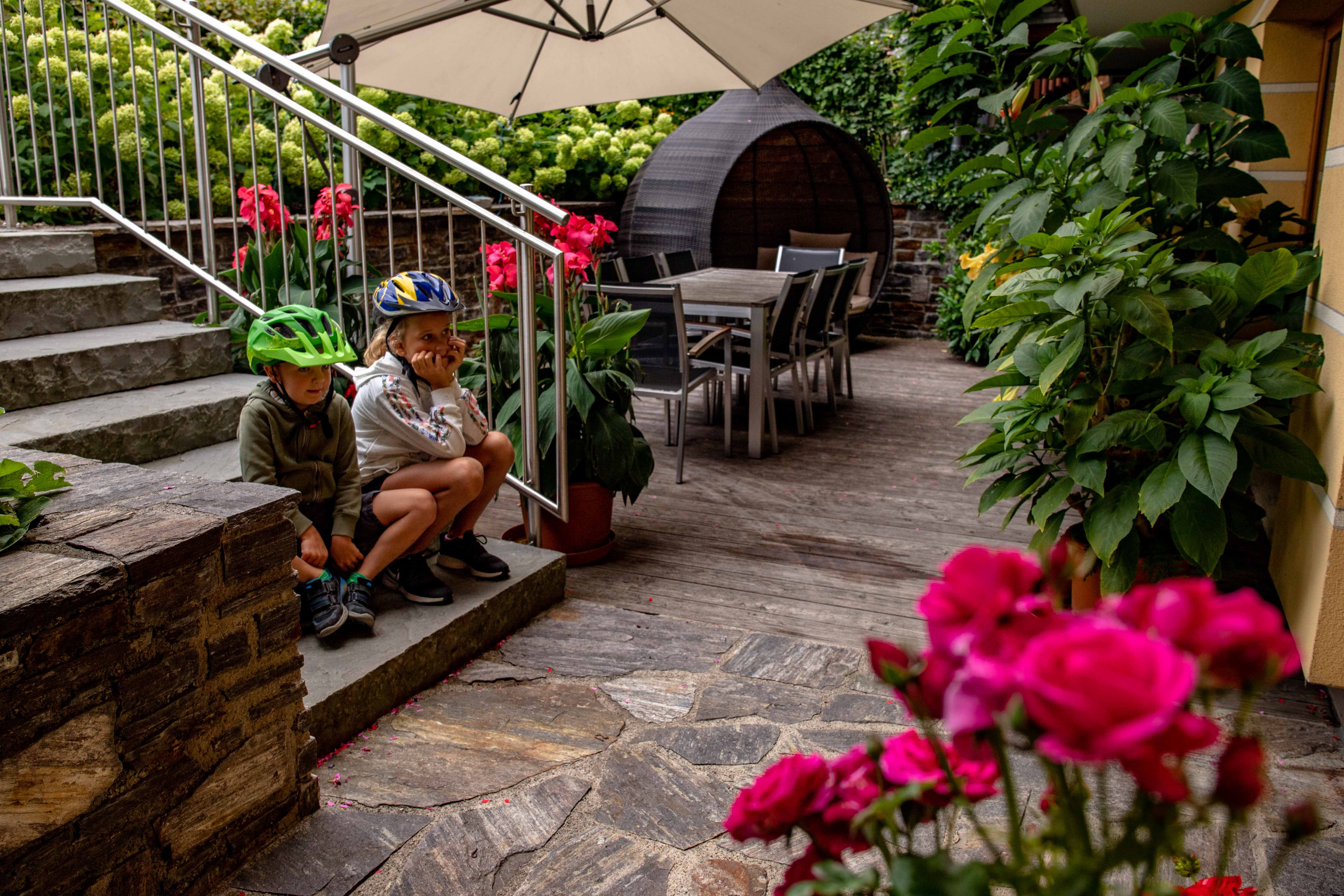 Two children wearing bicycle helmets are sitting on steps in a garden with flowers and garden furniture.