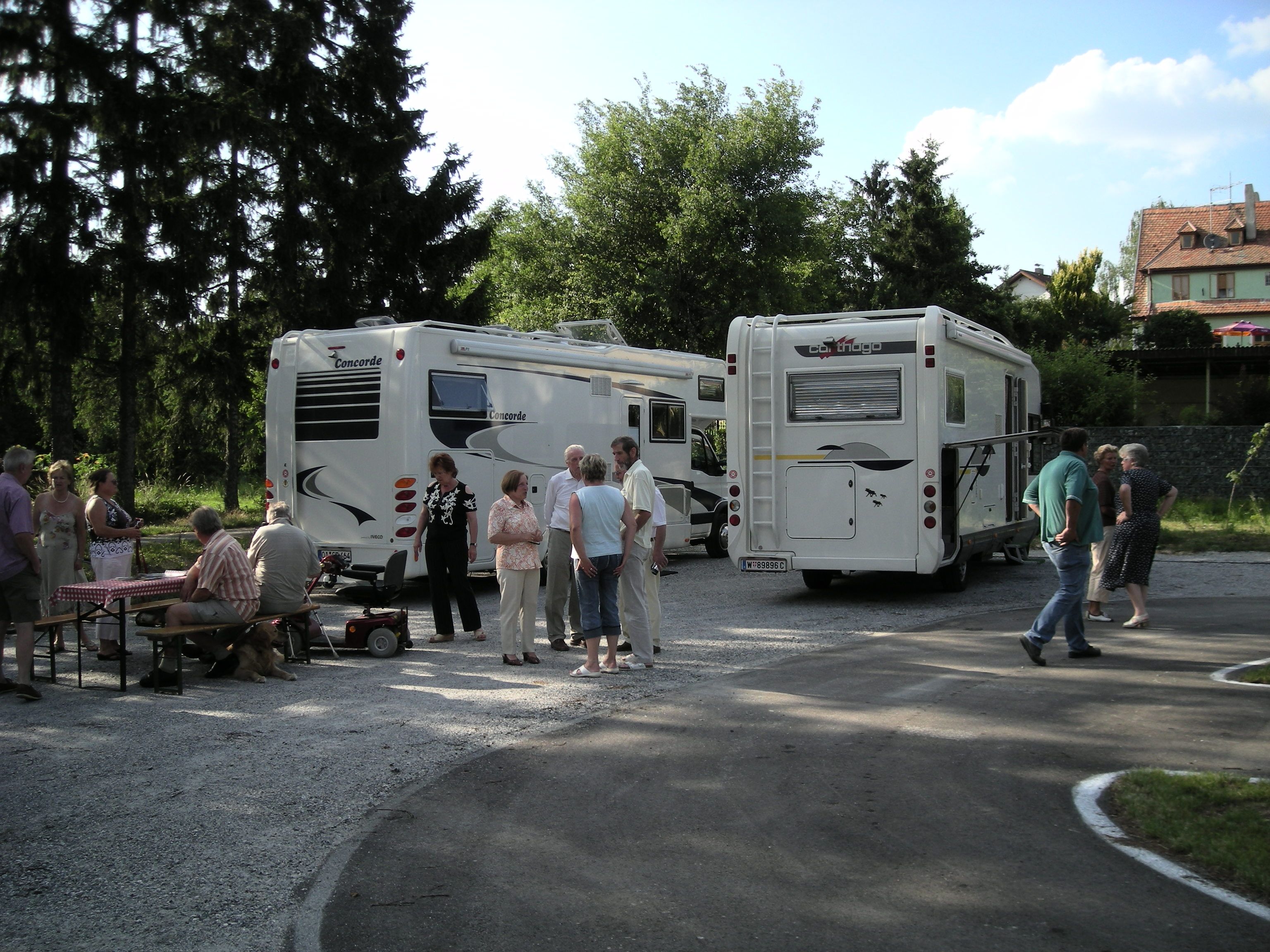 People stand and sit next to two mobile homes on an outdoor pitch.
