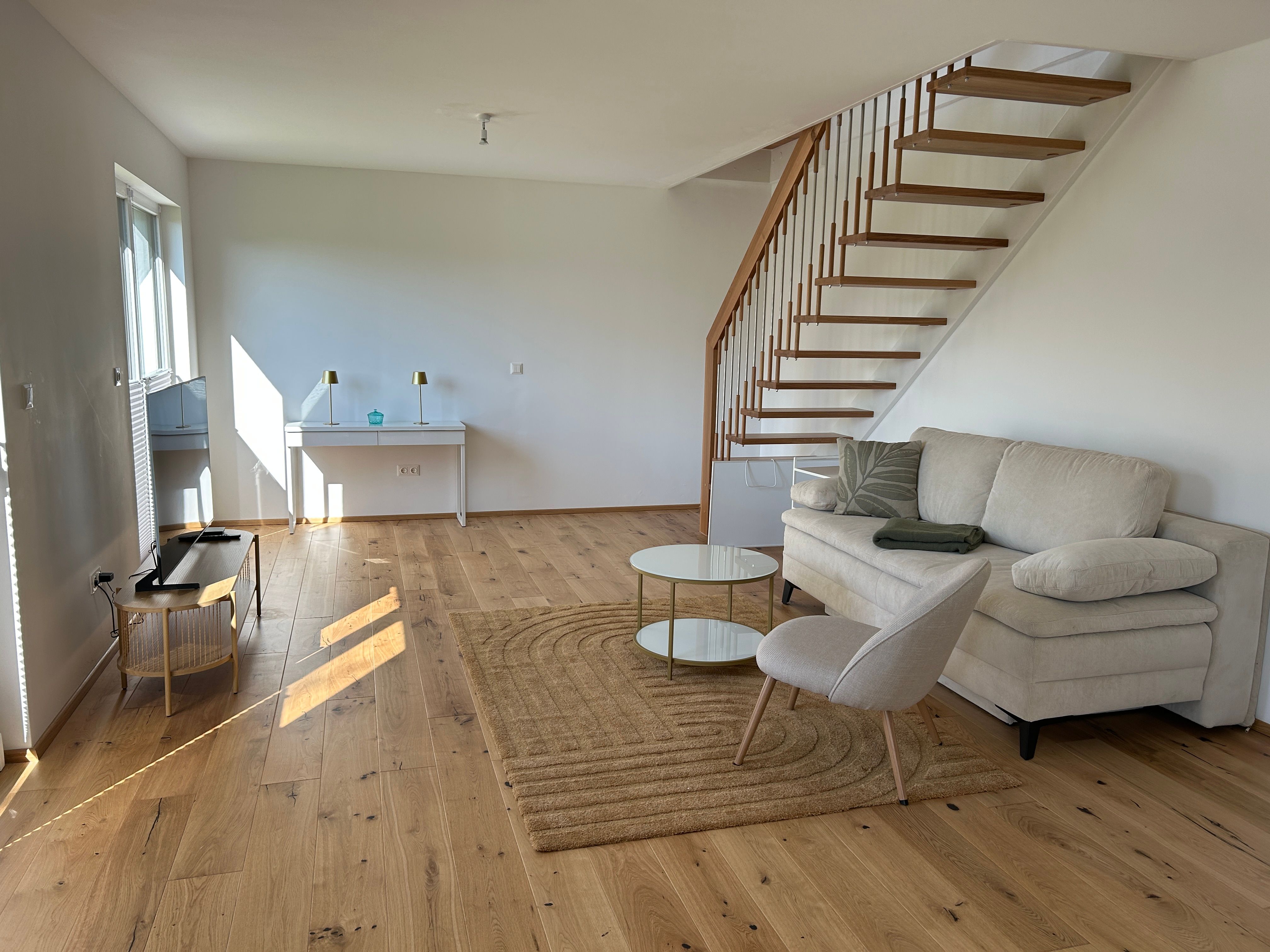 Modern living room with wooden staircase, sofa, armchair and coffee table on wooden floor.