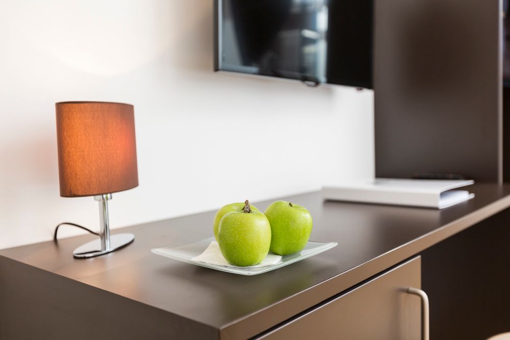 A table with three green apples on a plate, a lamp and a television in the background.