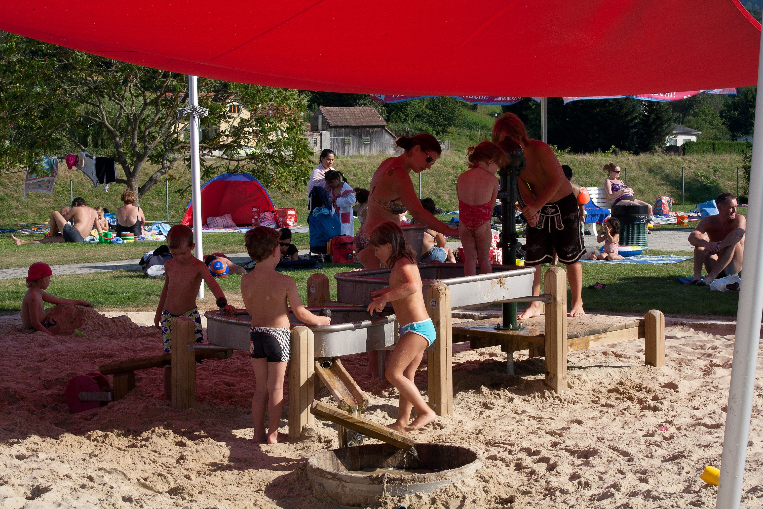 Children play in the sand under a red awning in Wilhelmsburg Parkbad.