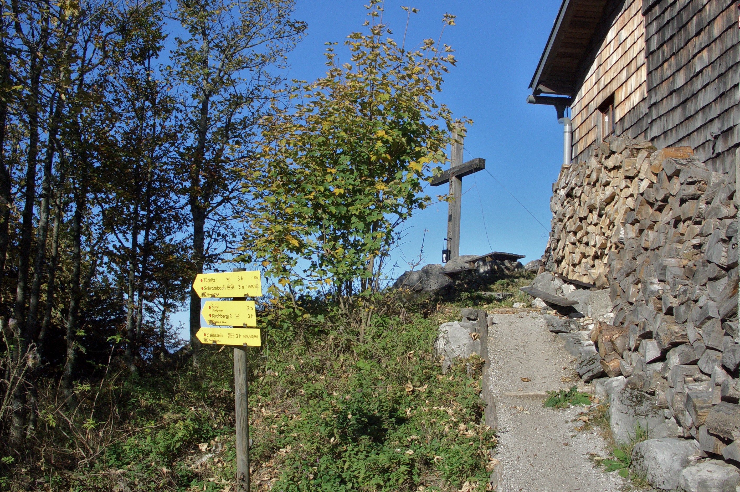 Signpost and wooden cross next to a wooden house in a mountainous landscape.