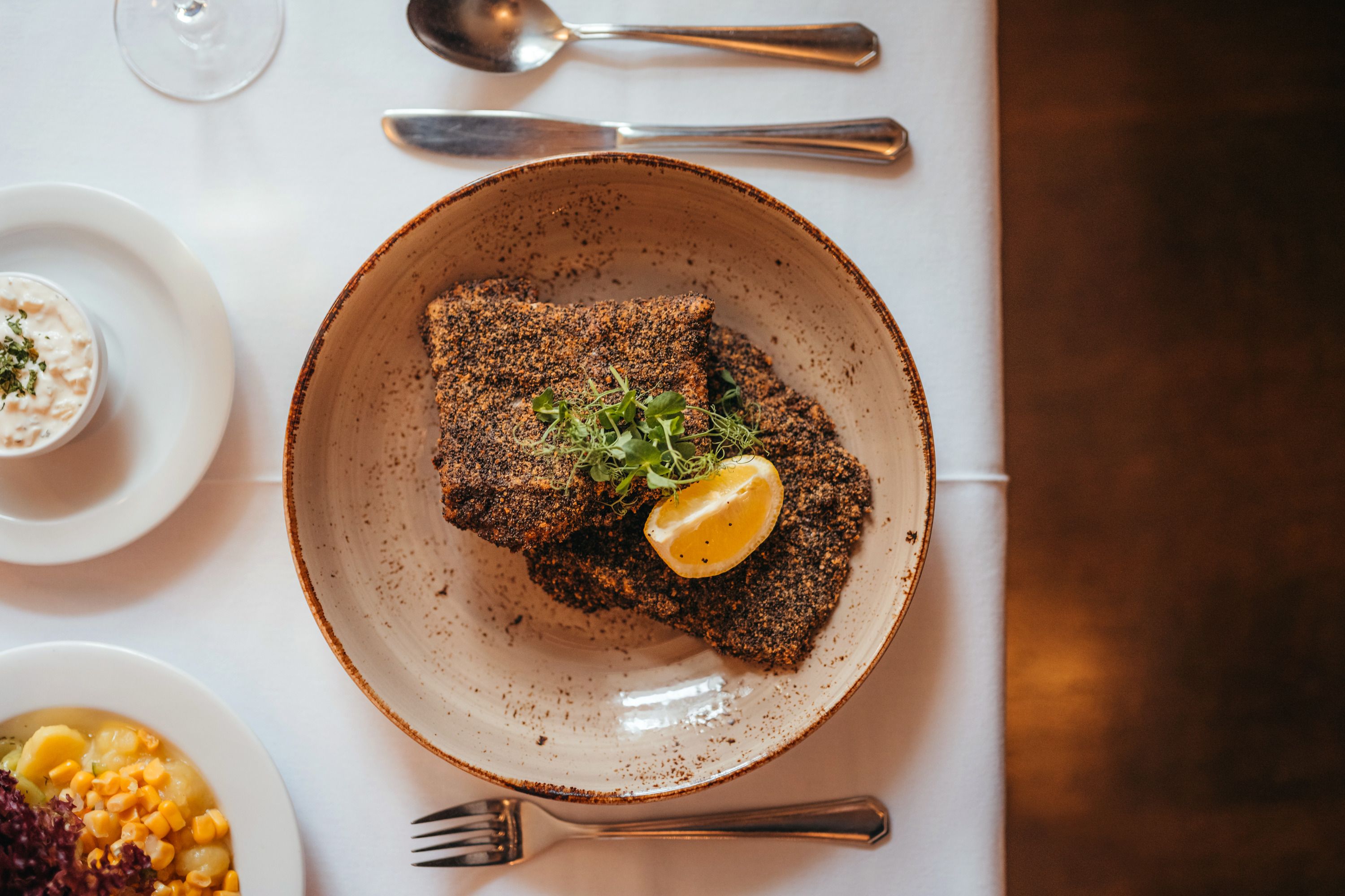 Grey poppy seed breaded carp fillet with lemon slice and herbs on a plate, side dishes next to it.