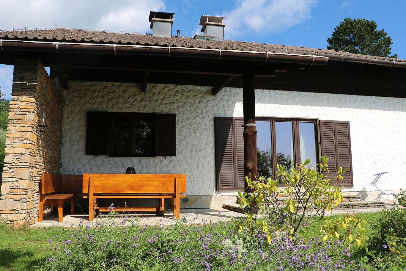 A house with a wooden seating area on the terrace, surrounded by a garden with flowers.