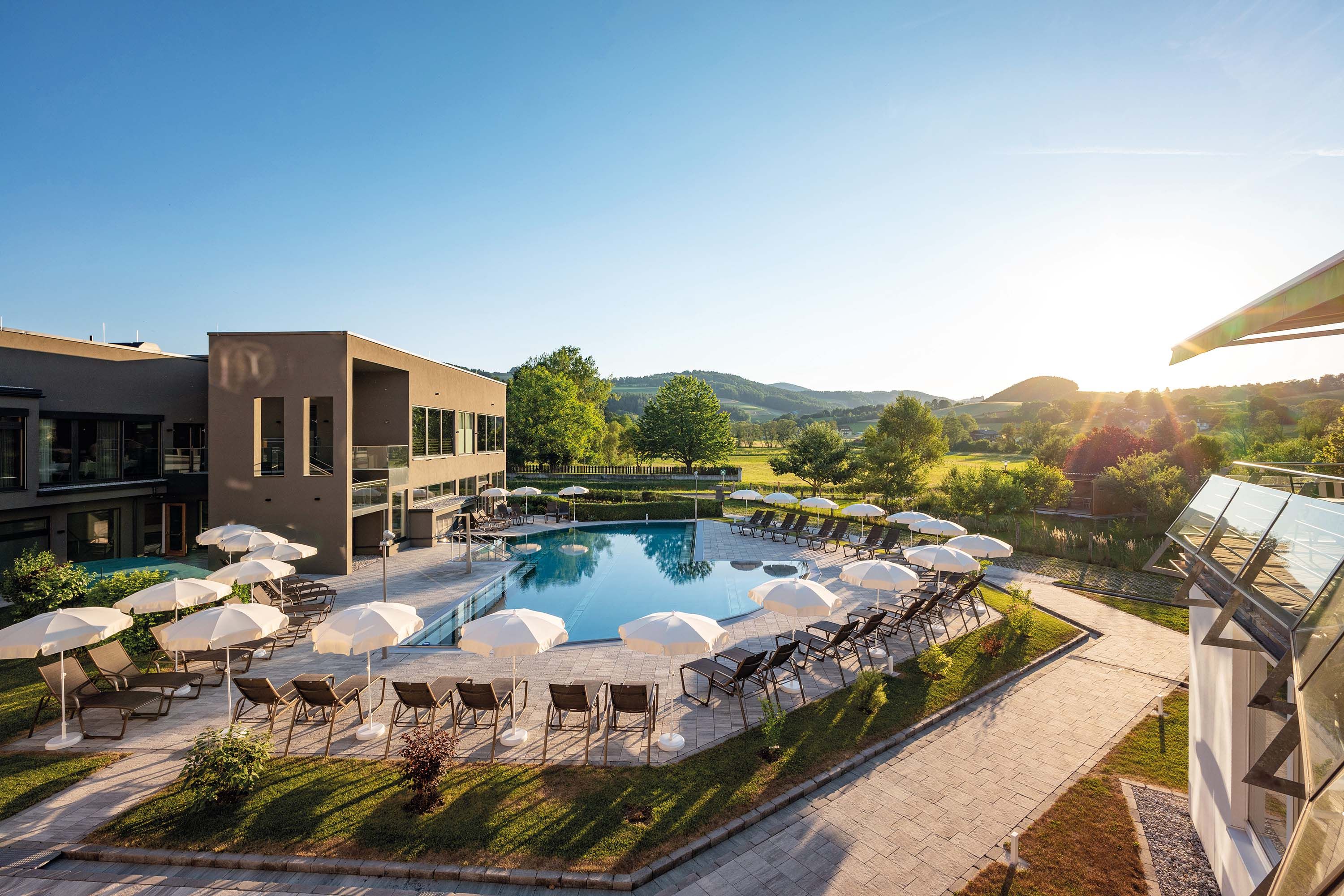 Outdoor area of a hotel with pool, parasols, sun loungers, surrounded by green meadows and trees.