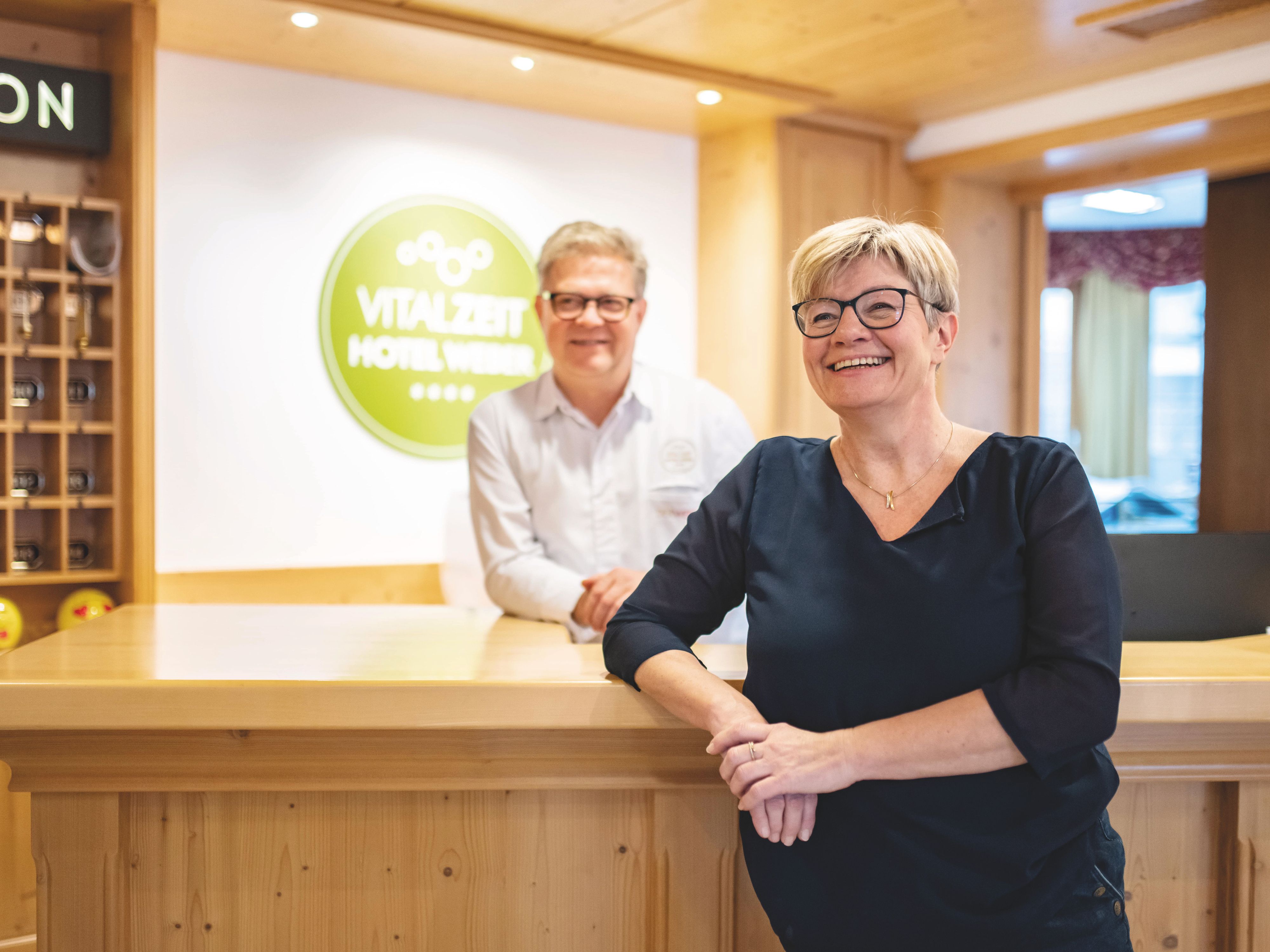 Petra and Alfred Weber are standing at the front desk of the VitalZeit Hotel Weber, smiling.