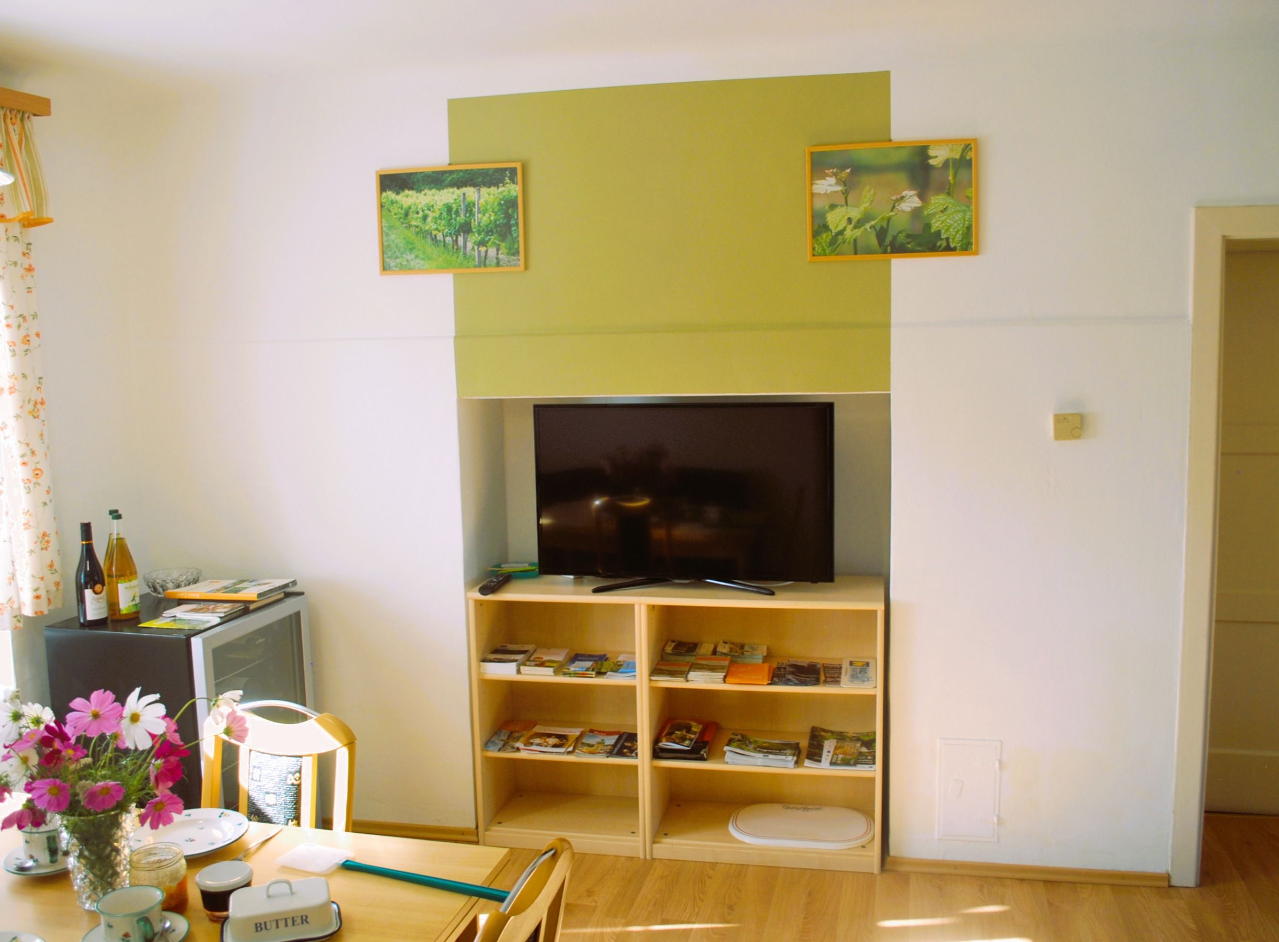A living room with a television on a shelf, surrounded by brochures. Two pictures of vineyards hang on the wall. A table with flowers and crockery is on the left.