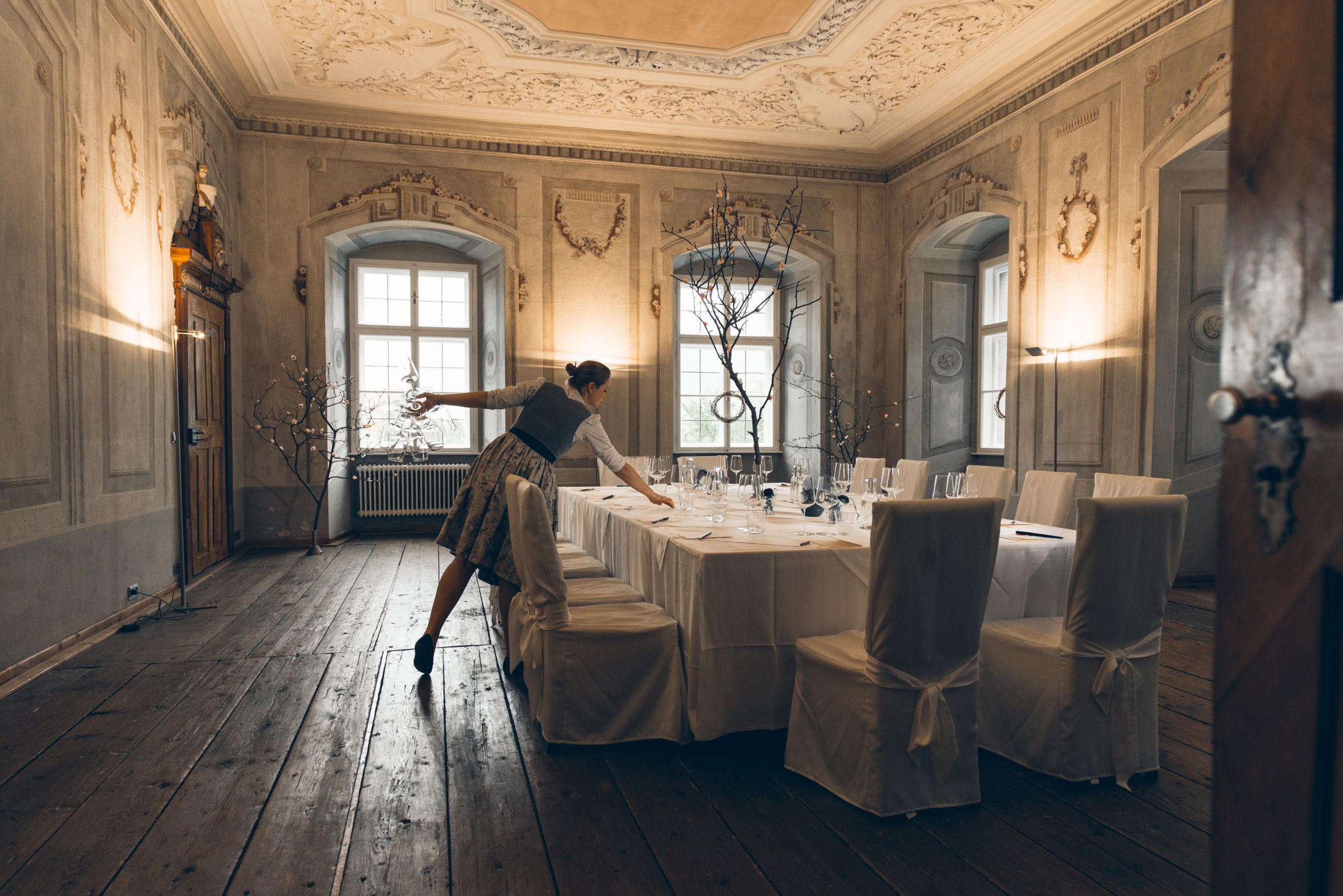 A woman prepares an elegantly laid table in a historic room with ornate walls and ceiling.