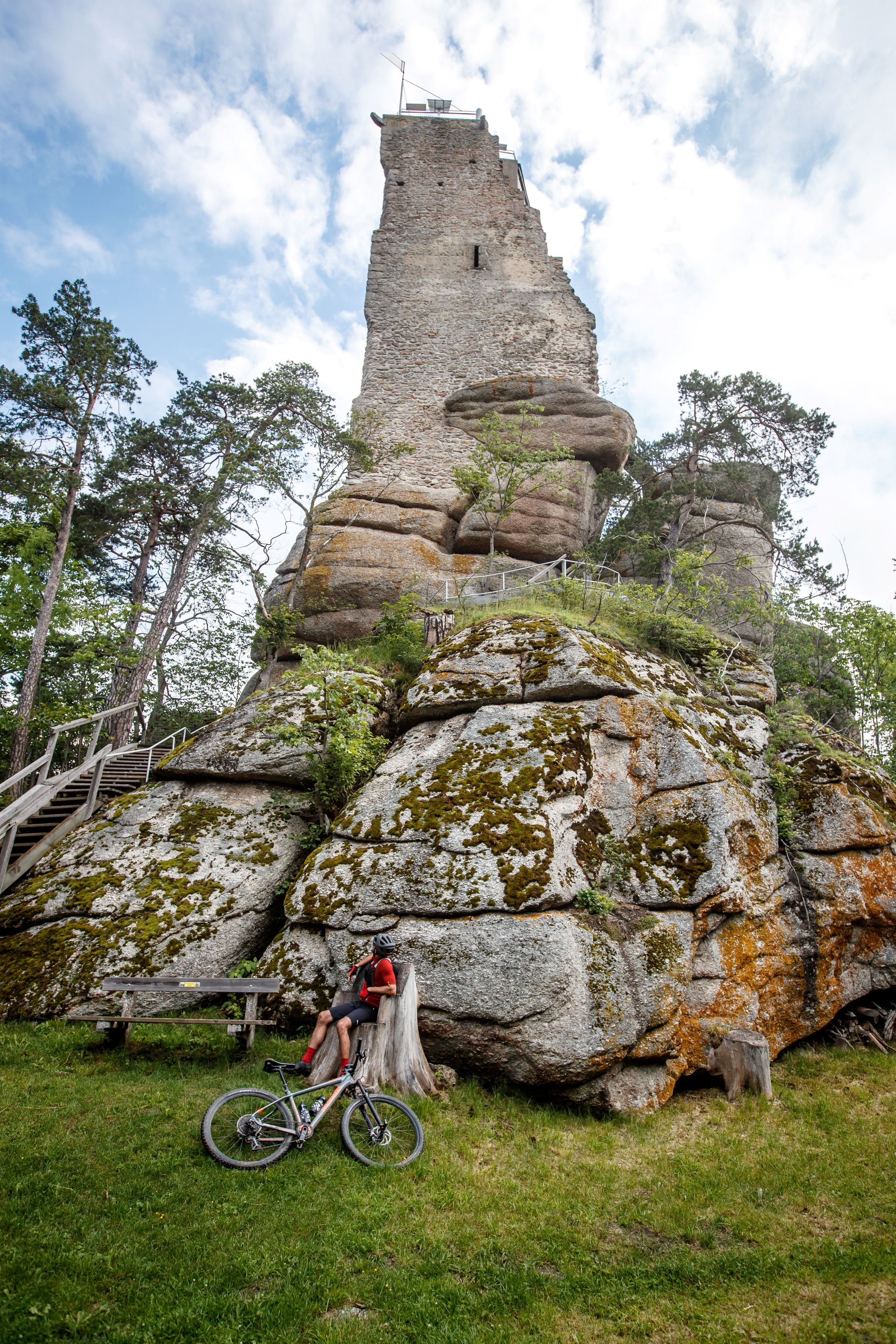 A cyclist leans against a tree stump in front of a high, rocky tower in a wooded area.