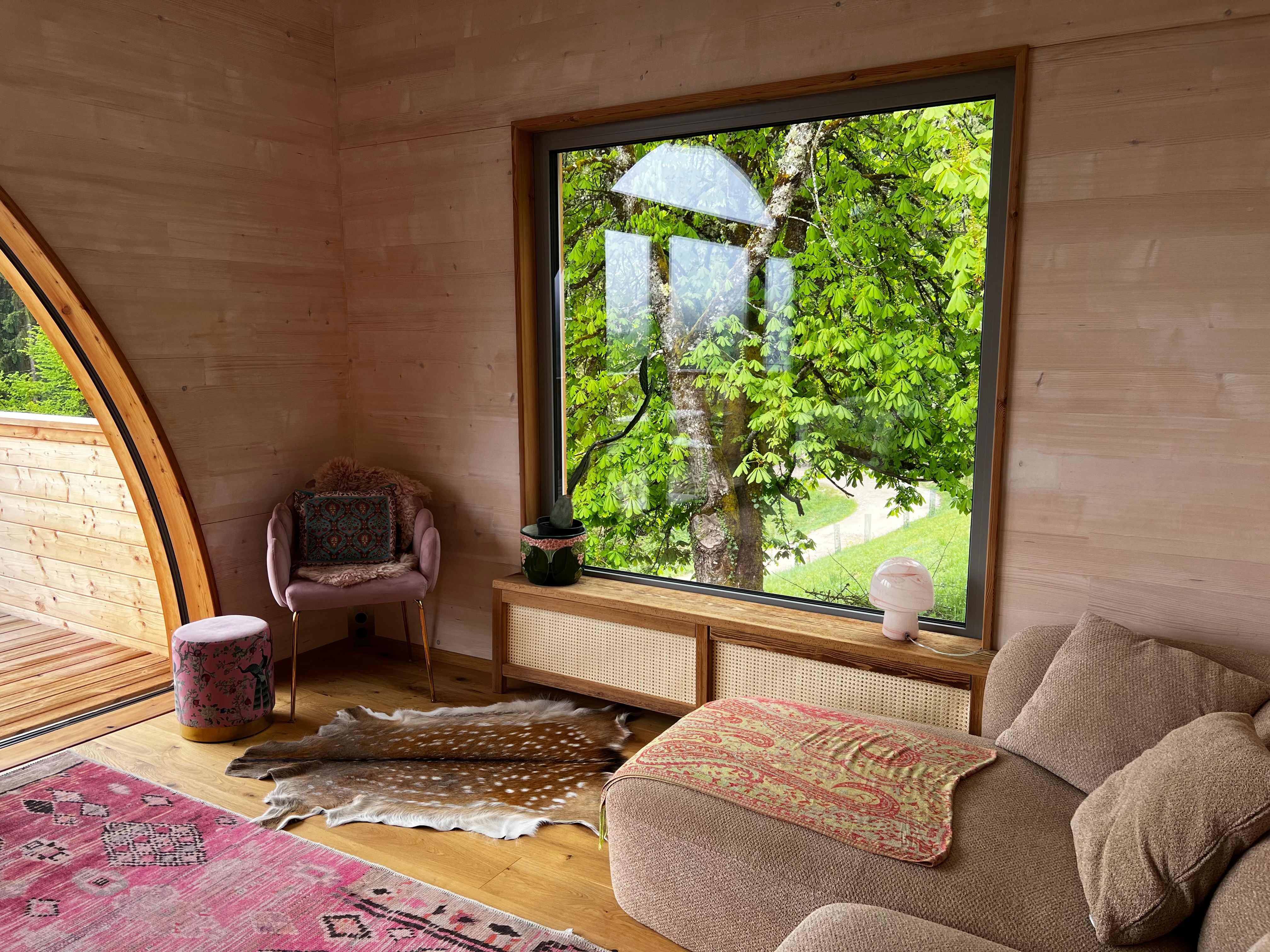 Interior view of a cozy room with a large window overlooking a green tree. An armchair, a stool and a sofa can be seen.