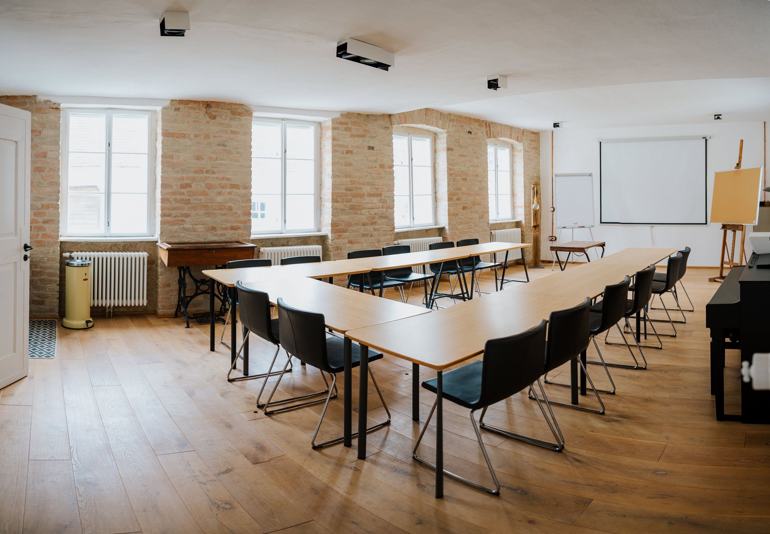 A bright seminar room with a U-shaped table arrangement, chairs, projection screen and flipchart.