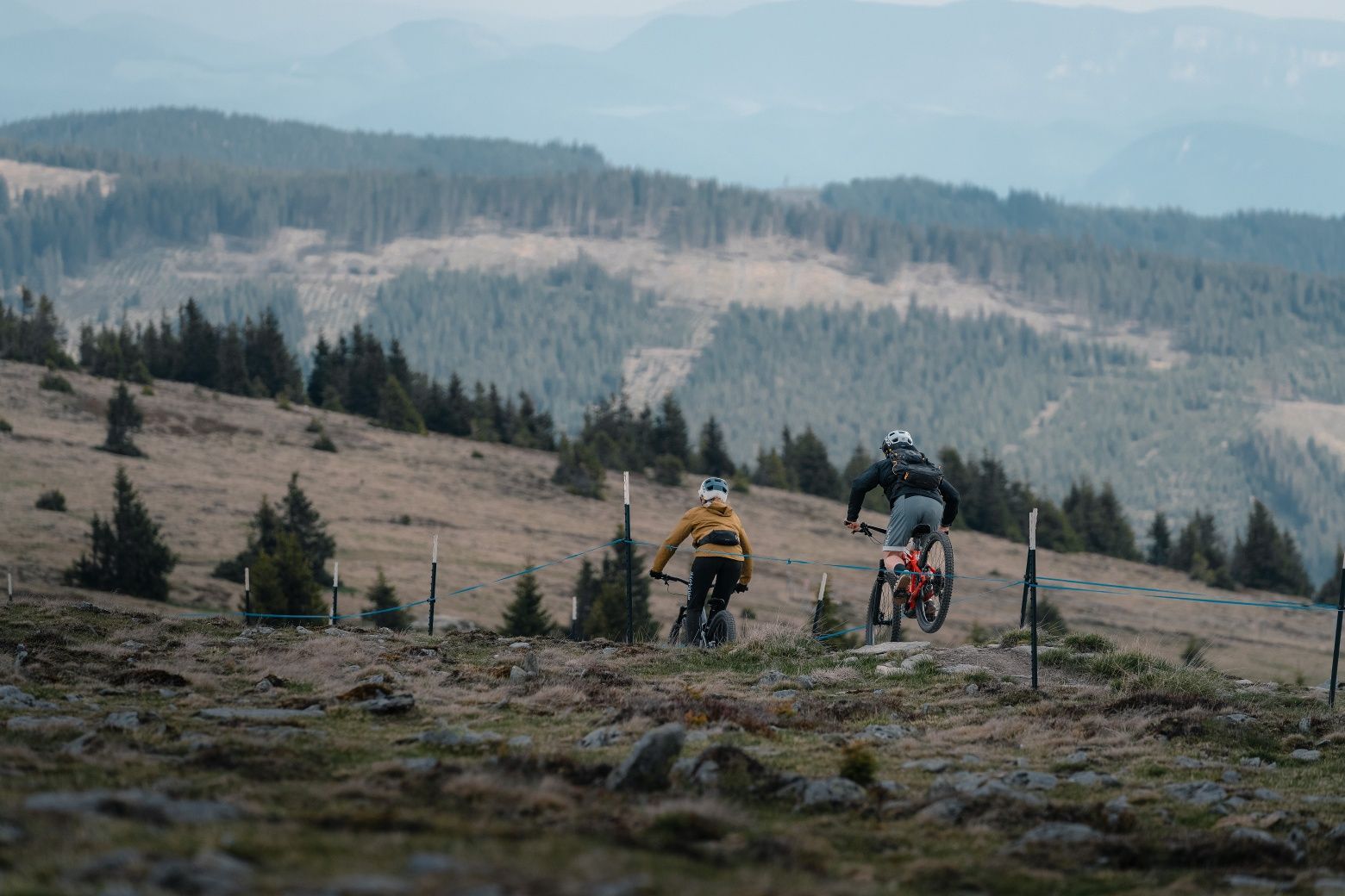 Two mountain bikers ride down a path in the mountains, surrounded by forests and hills.