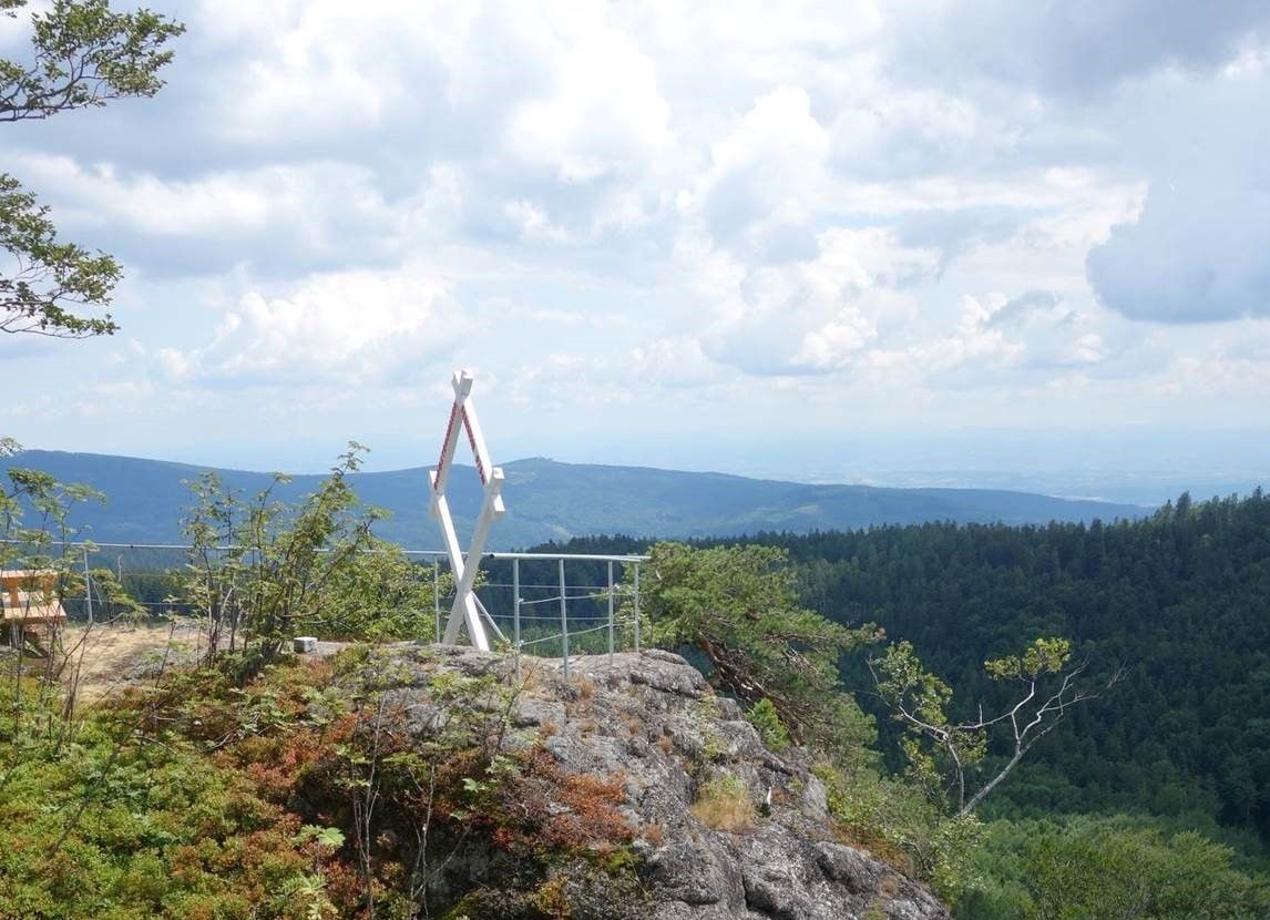 Viewpoint on a mountain with a white, cross-shaped frame and a view of wooded hills under a cloudy sky.