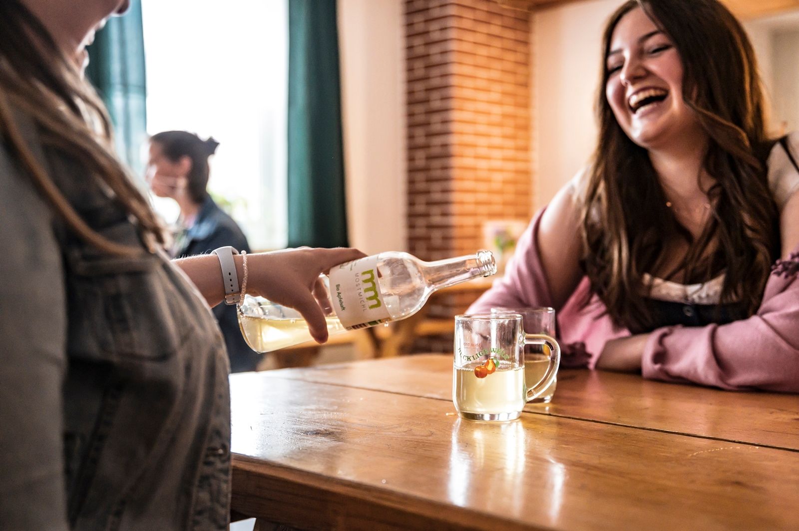 Two women are sitting at a table, one is pouring Cider from a bottle into a glass. They are both laughing.