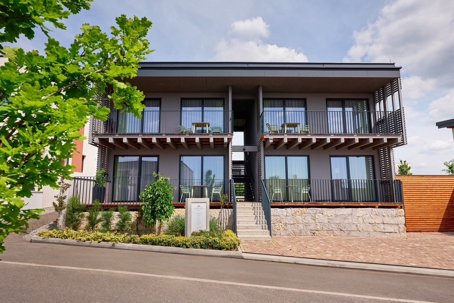 Modern two-storey apartment complex with balconies and staircase, surrounded by plants and trees.
