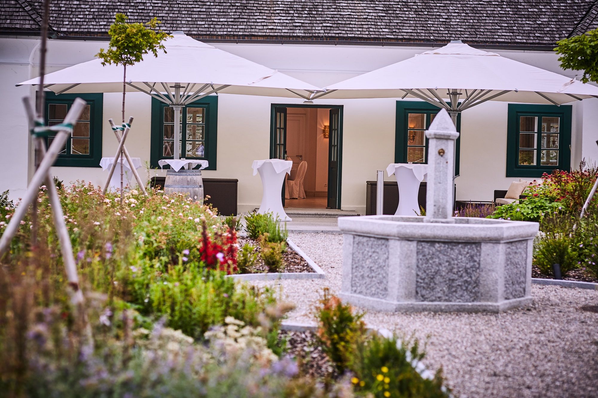 A stone fountain in a garden in front of a building with white sunshades and green shutters.