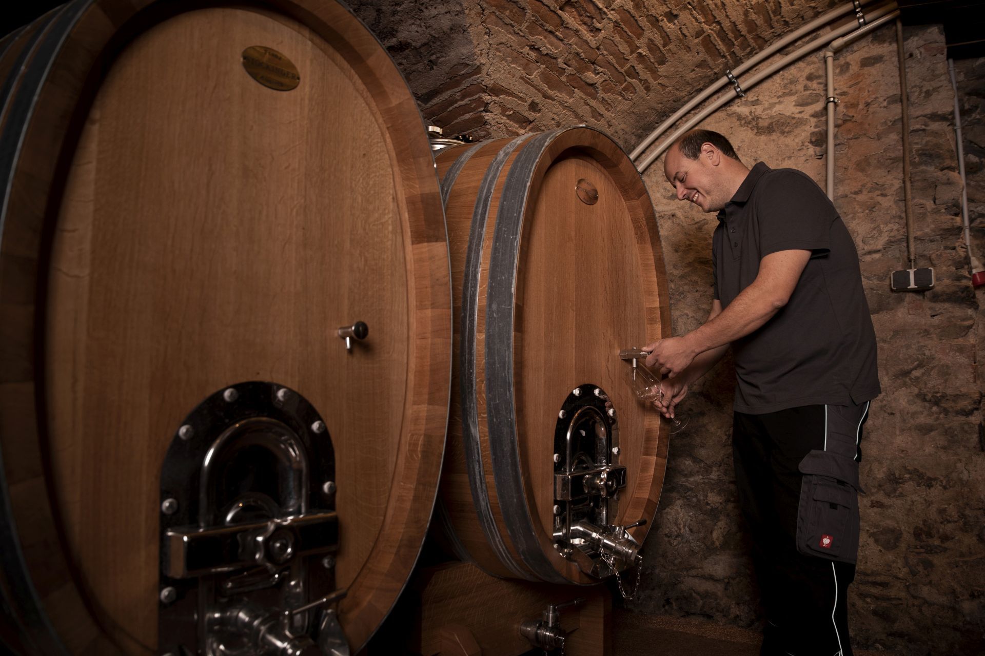 A man draws wine from a large wooden barrel in a wine cellar.