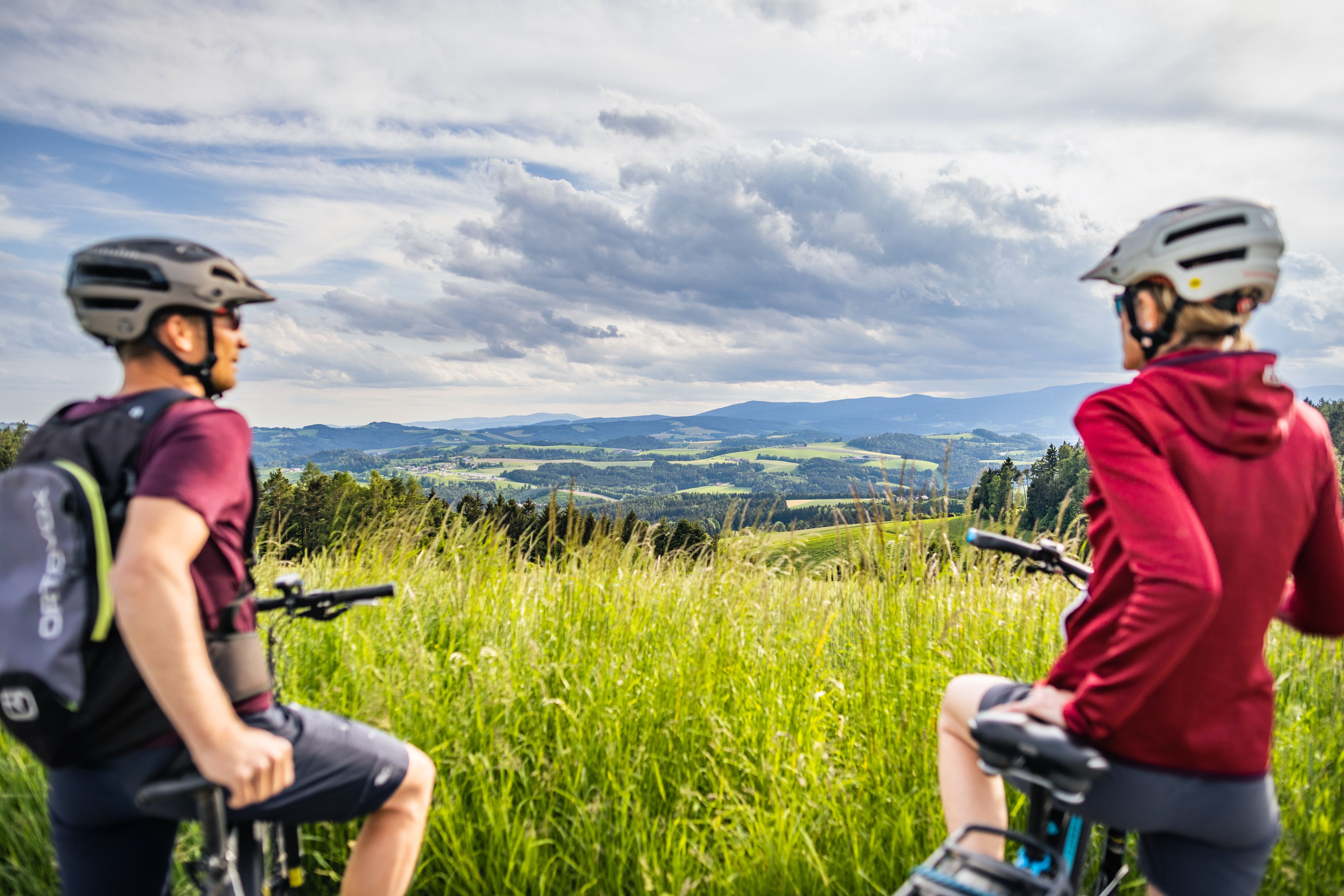 Two cyclists with helmets look out over a hilly landscape of meadows and forests under a cloudy sky.