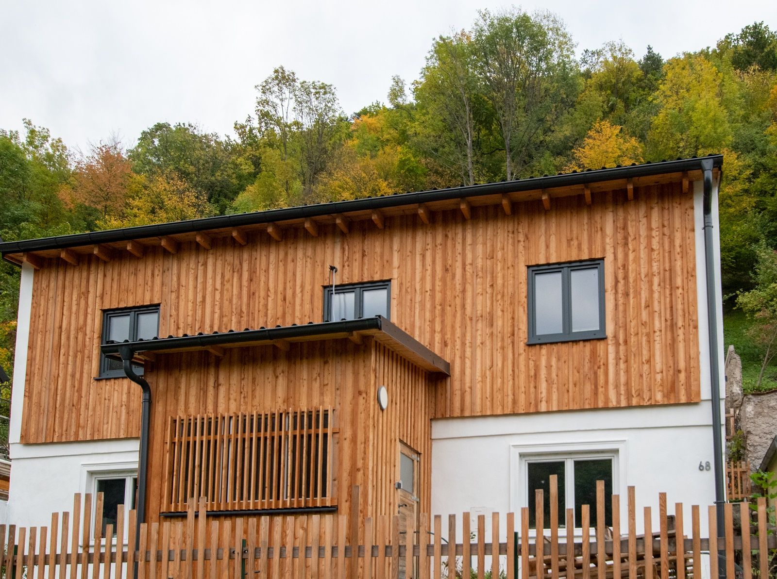 A two-story wooden house with modern windows in front of a wooded hill.