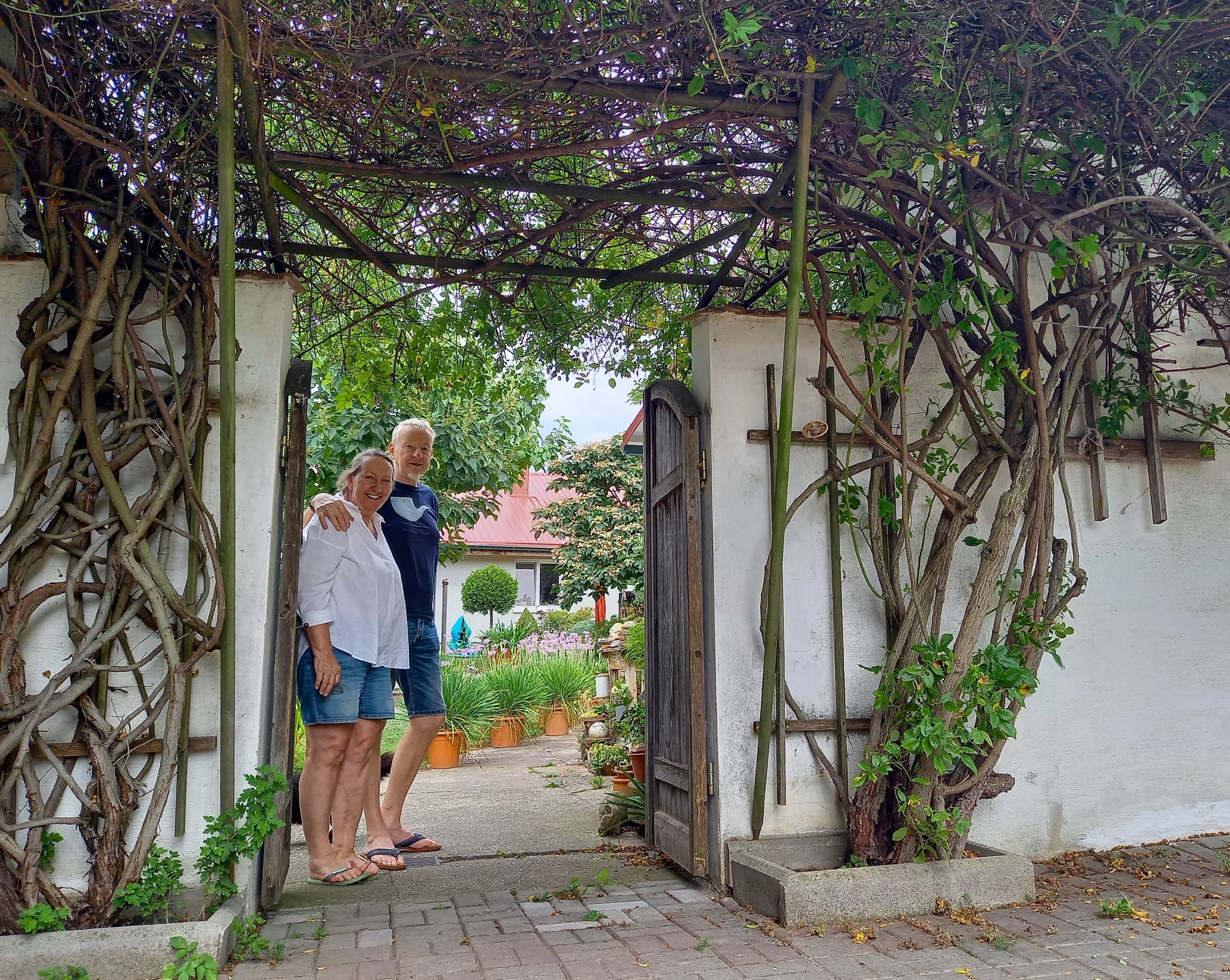 Two people stand smiling under an archway overgrown with plants in a garden.