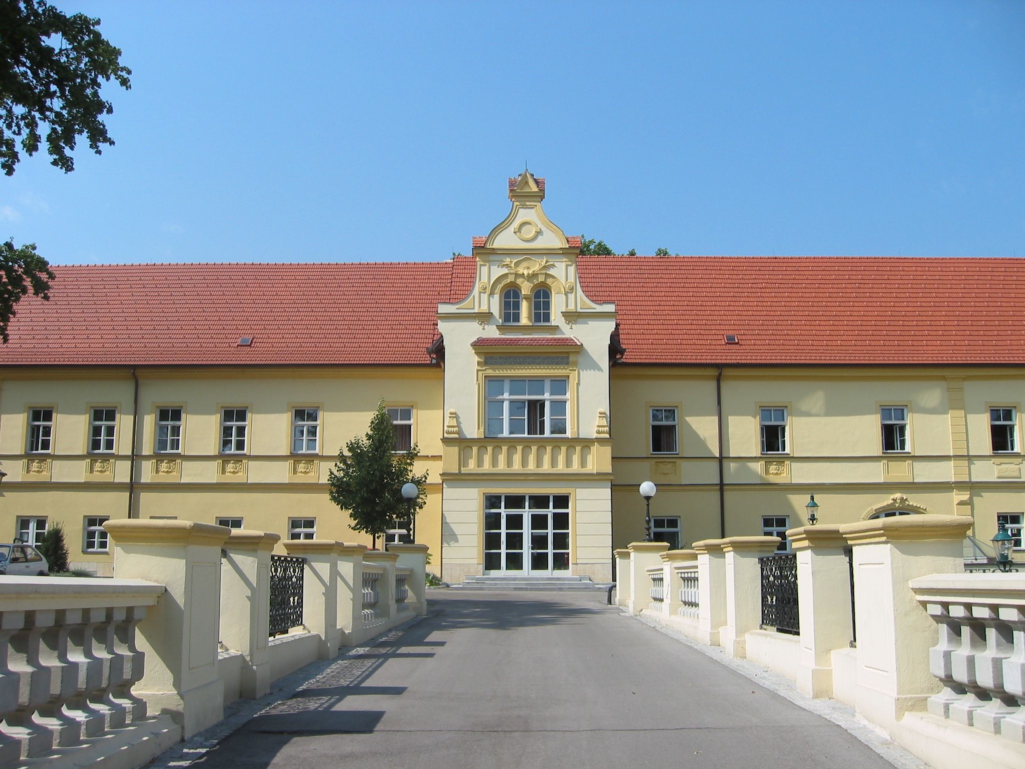 Yellow building with a red roof and decorative façade, surrounded by a bridge and trees.