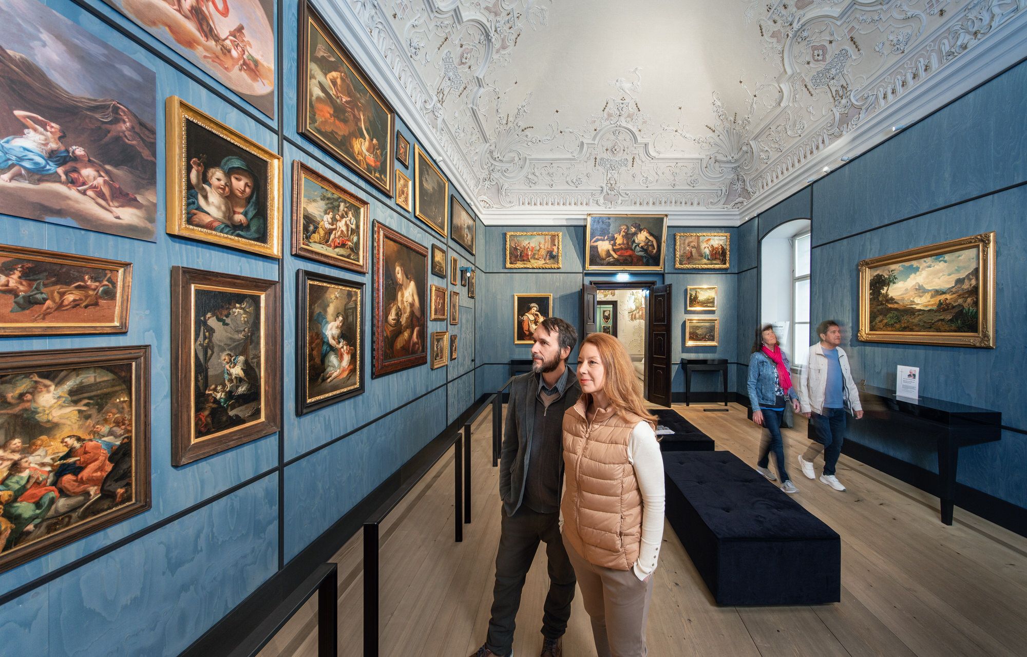 Visitors view paintings in an artistically decorated room with blue walls and ornate ceiling stucco.