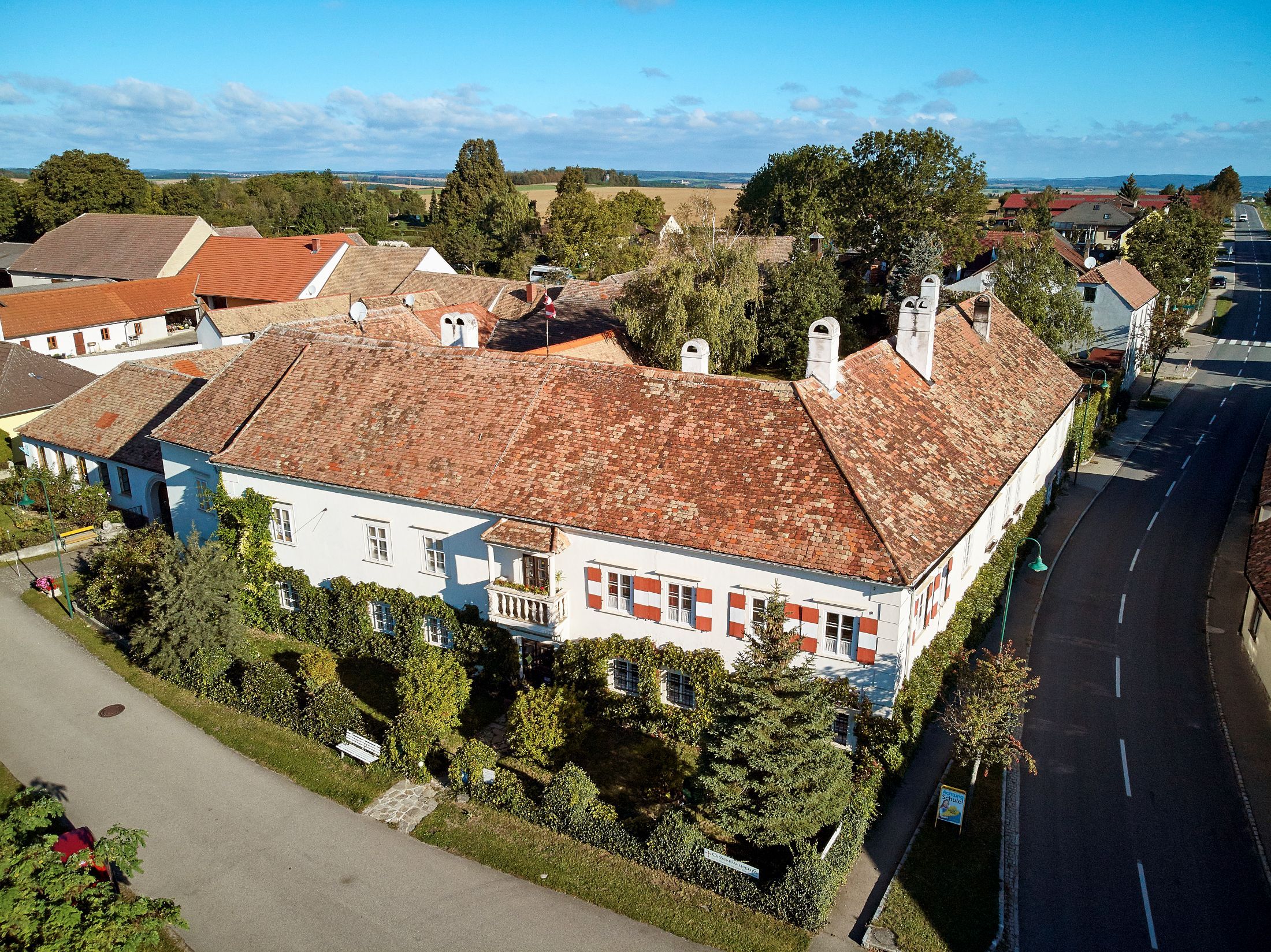 Aerial view of a historic building with red tiled roofs and white facades, surrounded by trees and streets.