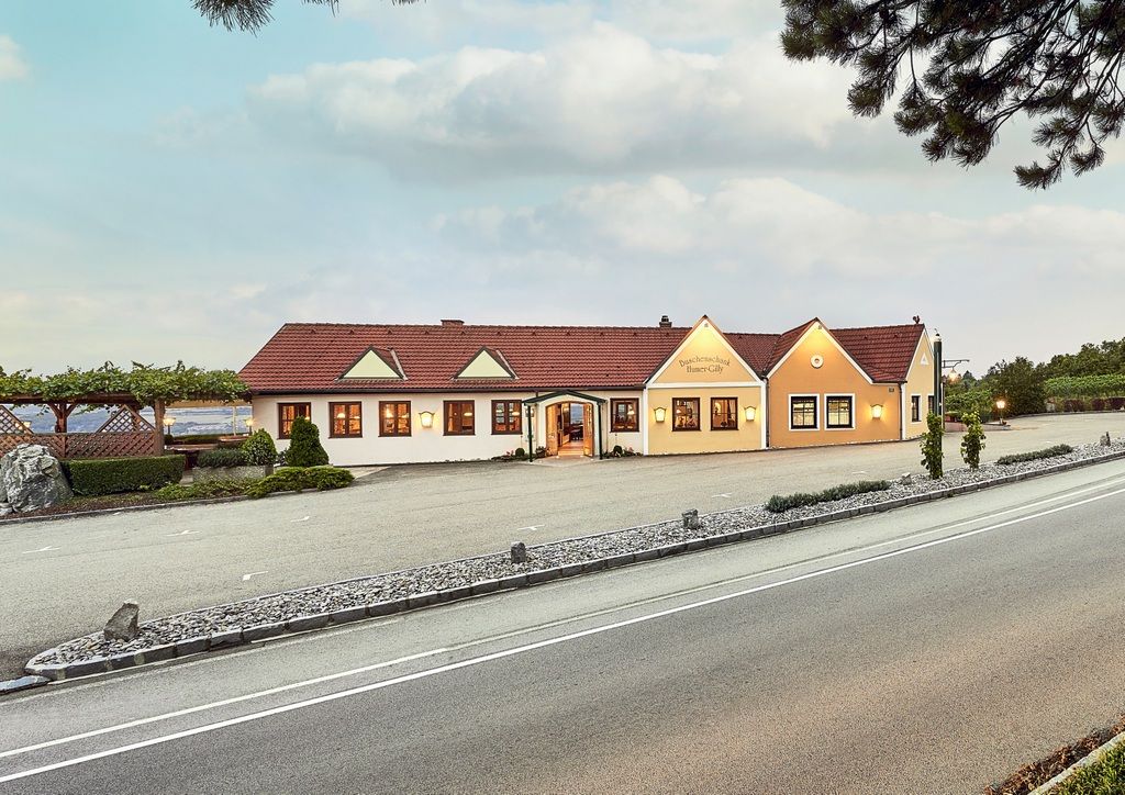Exterior view of a winery with a yellow building and red roof, surrounded by vines and a road in the foreground.