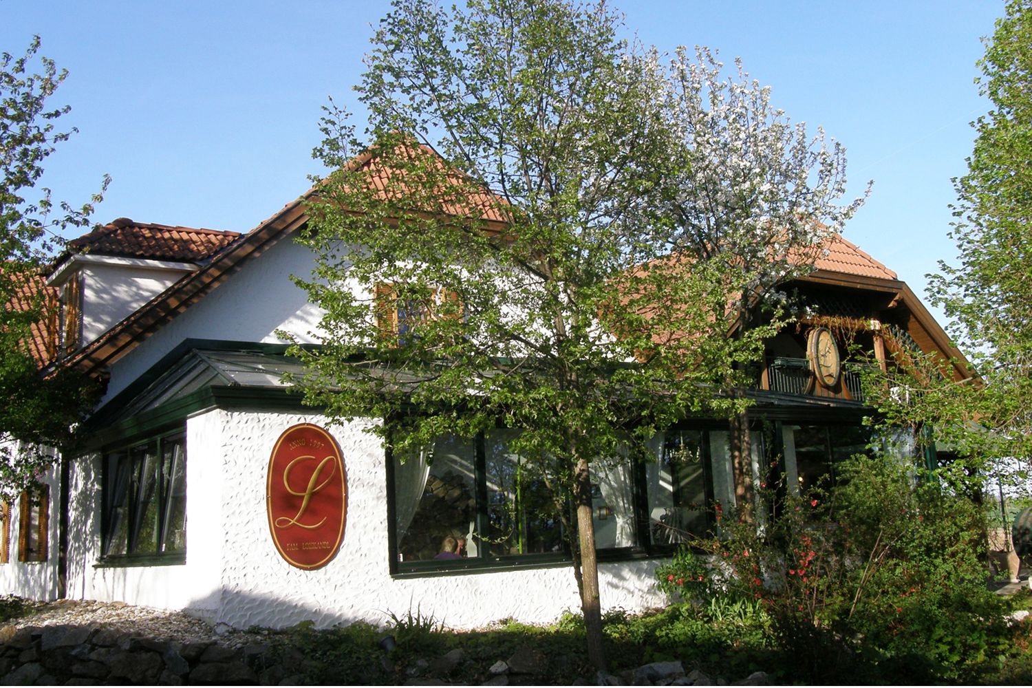 Exterior view of a traditional building with red roof tiles and a large L logo on the wall, surrounded by trees and plants.