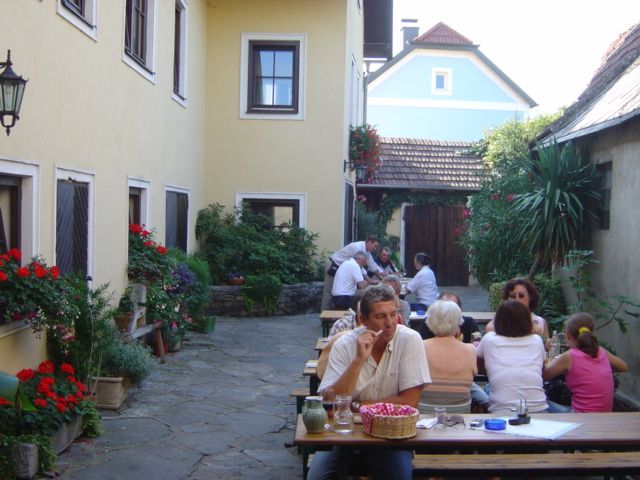 People sit at wooden tables in a courtyard with yellow buildings and plants.