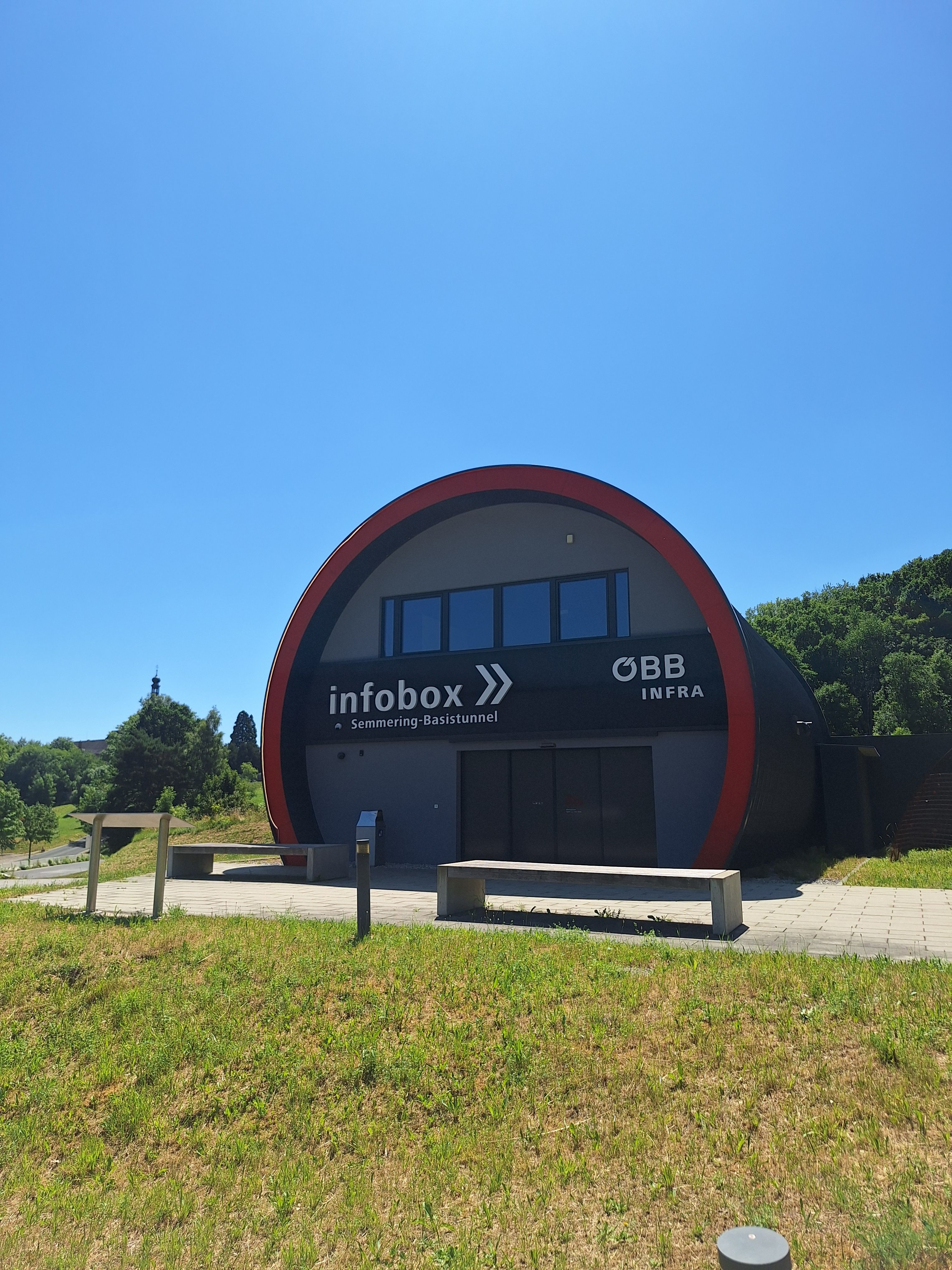 Building of the Semmering Base Tunnel info box with ÖBB logo, surrounded by green meadow and trees, under a blue sky.