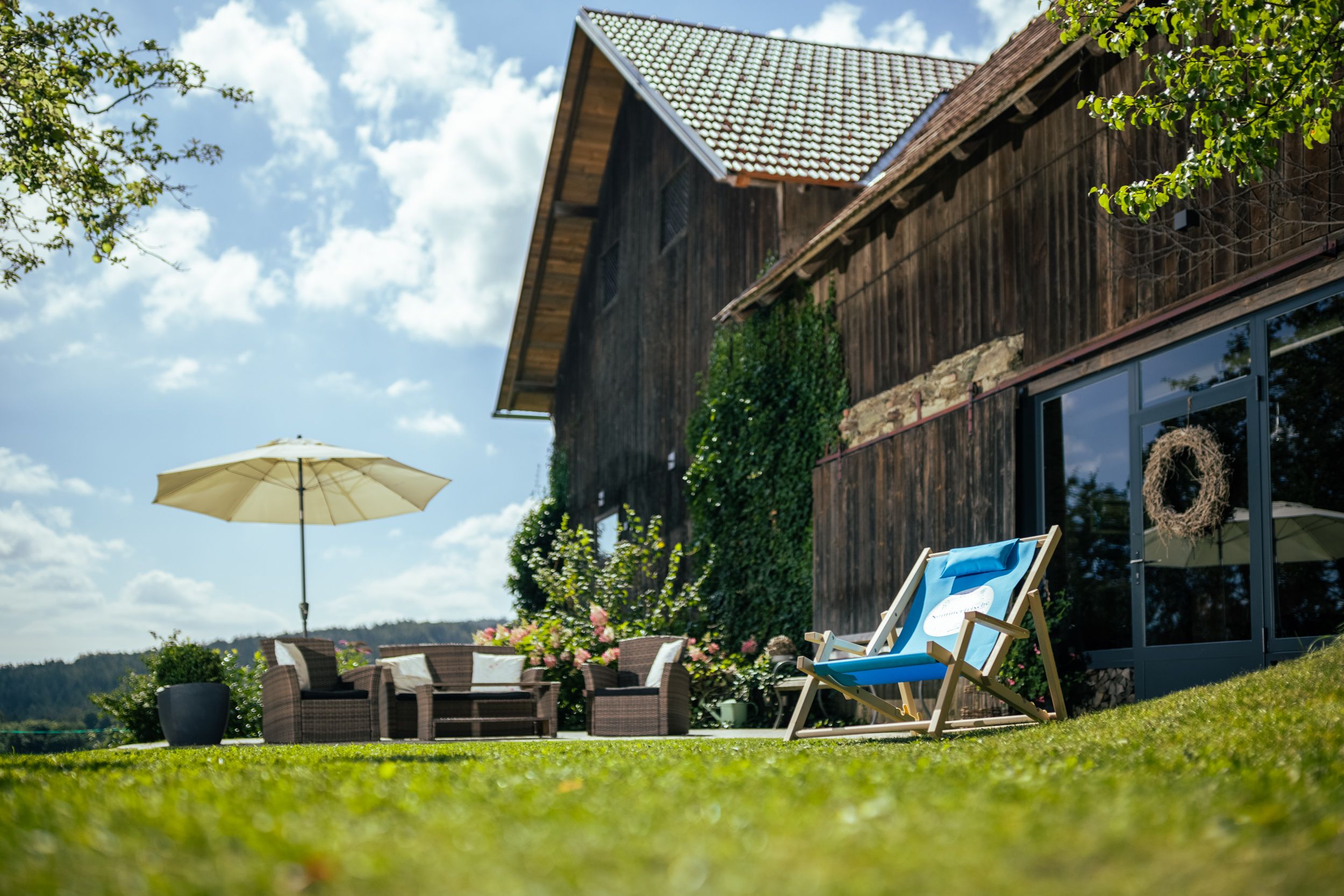 A farmhouse with a wooden façade, garden furniture and parasol on a meadow in sunny weather.