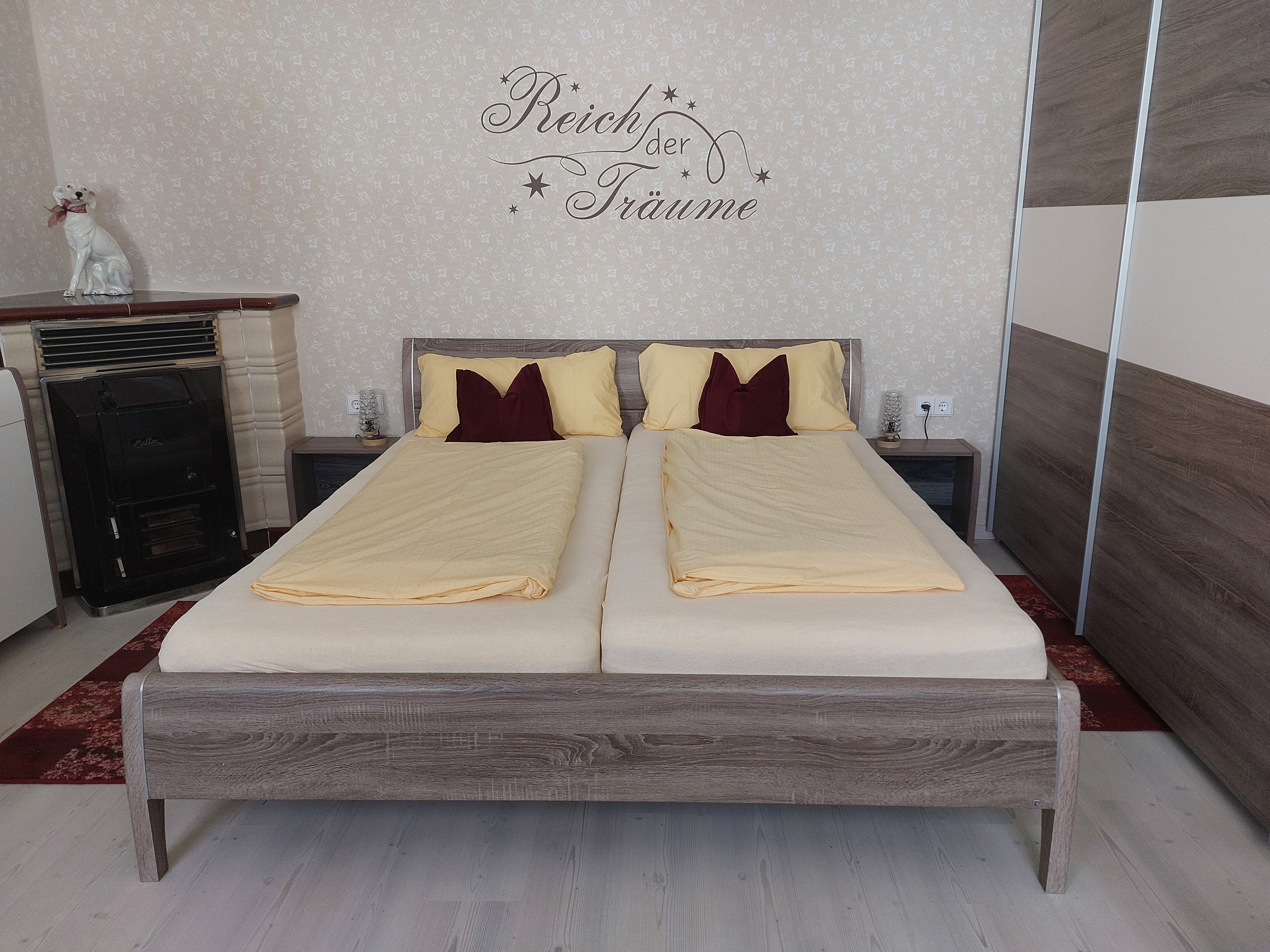 A bedroom with a double bed, yellow bed linen and writing on the wall. A stove and a dog sculpture can also be seen.