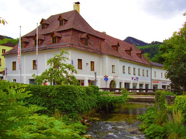 Historic building with a red roof by a stream, surrounded by green vegetation.