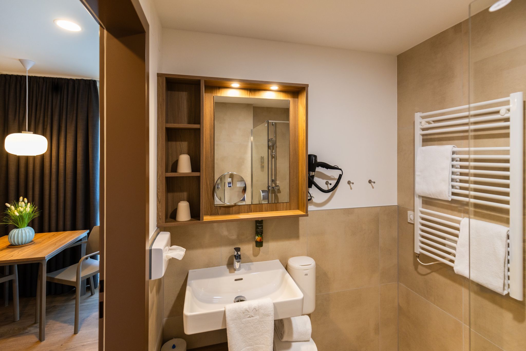 A modern bathroom with a sink, mirror, towel rack, and a view into an adjoining room with a table and a vase.