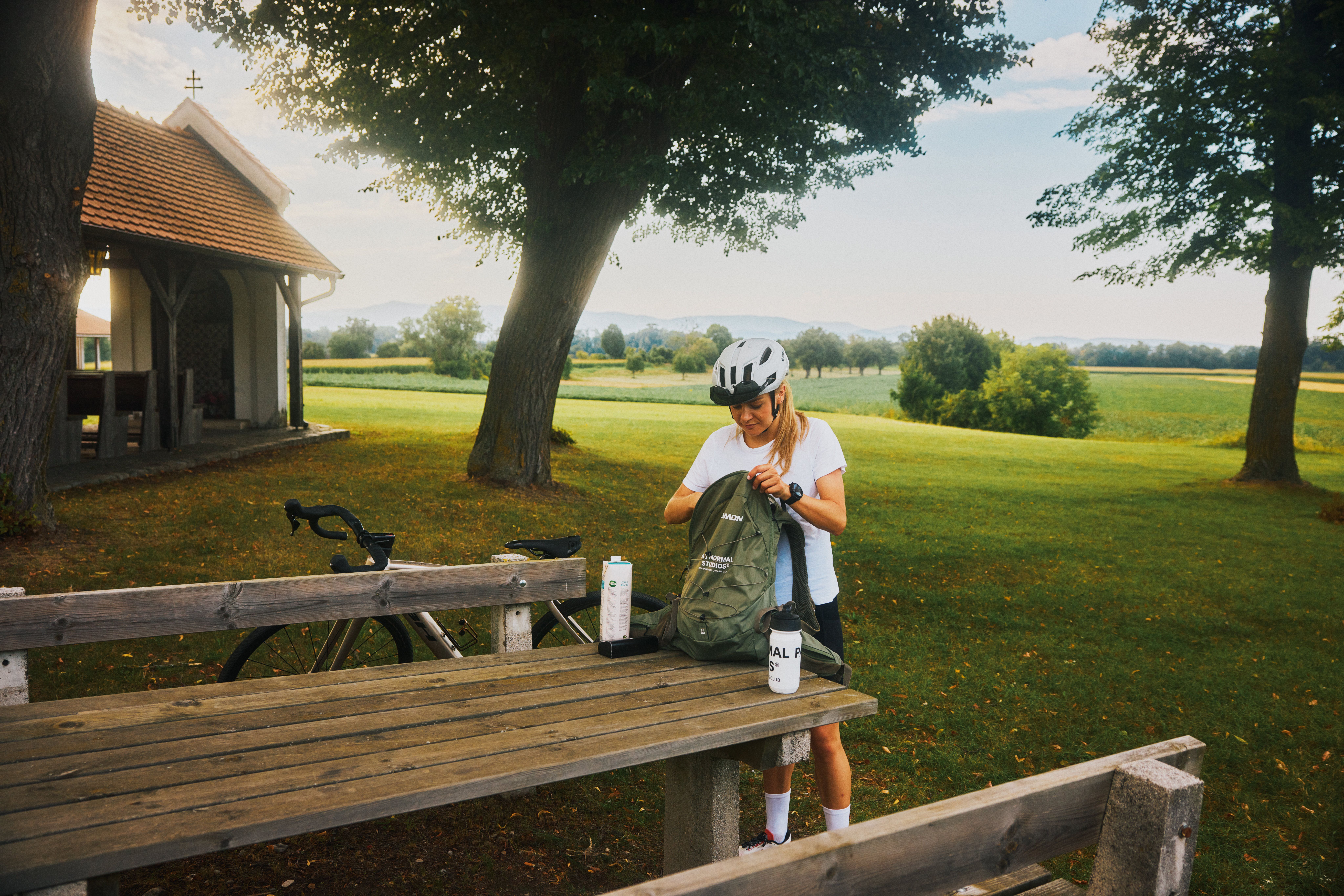 A woman with a bicycle helmet stands at a picnic table in a rural setting and packs her rucksack. A small chapel can be seen in the background.